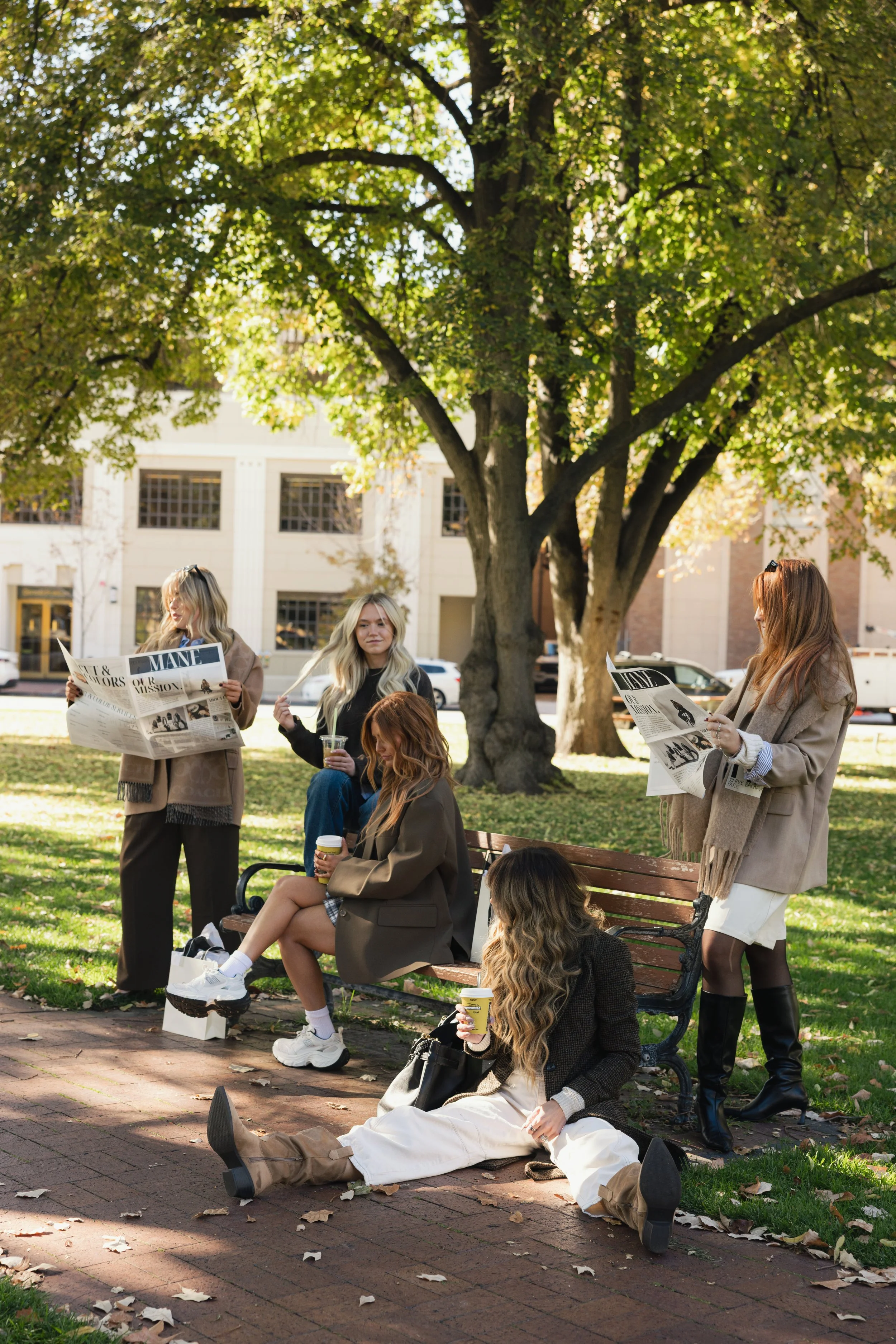 Five women in autumn outfits relaxing and reading newspapers in a park on a sunny day, with some sitting on a bench and others standing nearby.