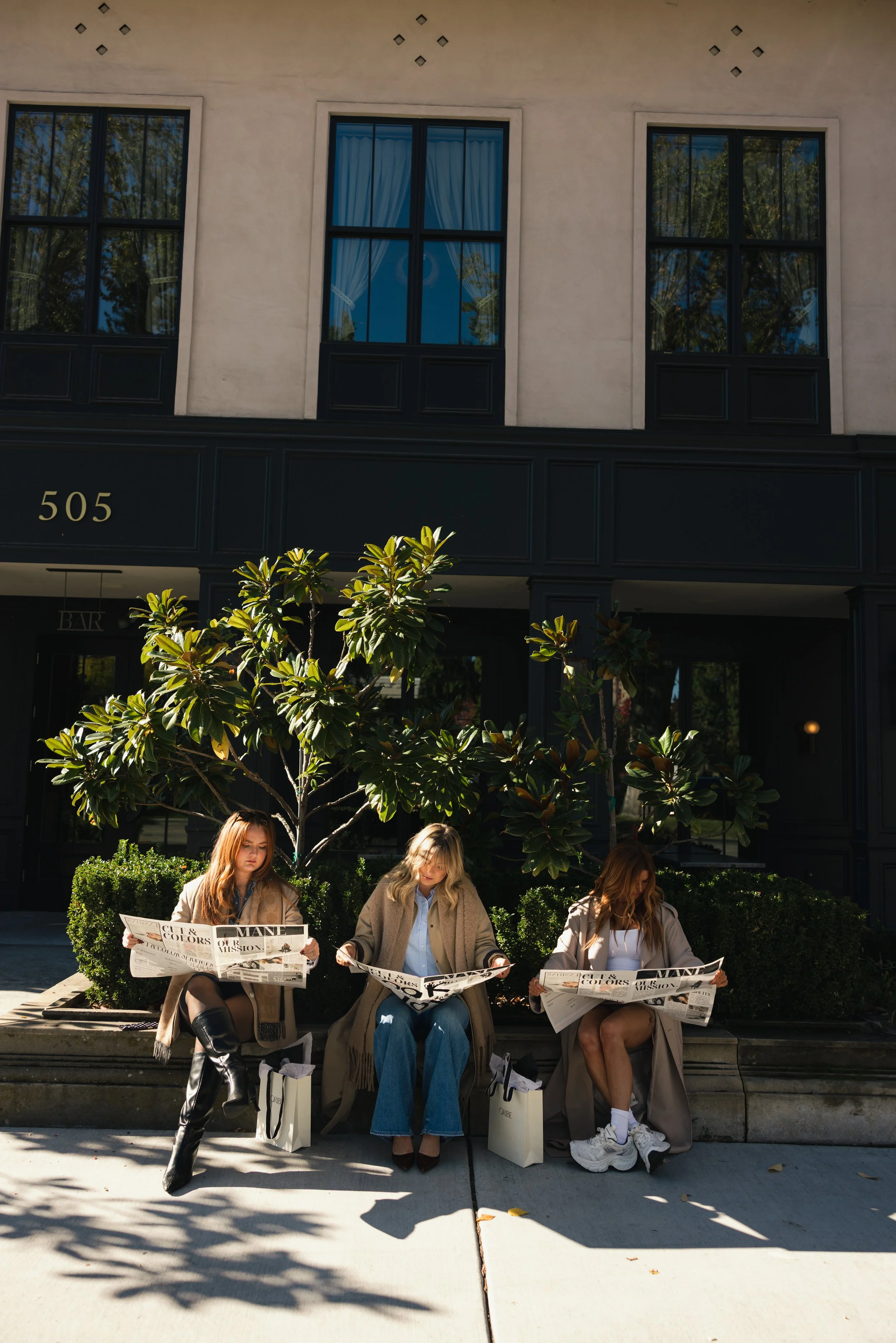 Three women sitting on a sidewalk outside a building, reading newspapers.