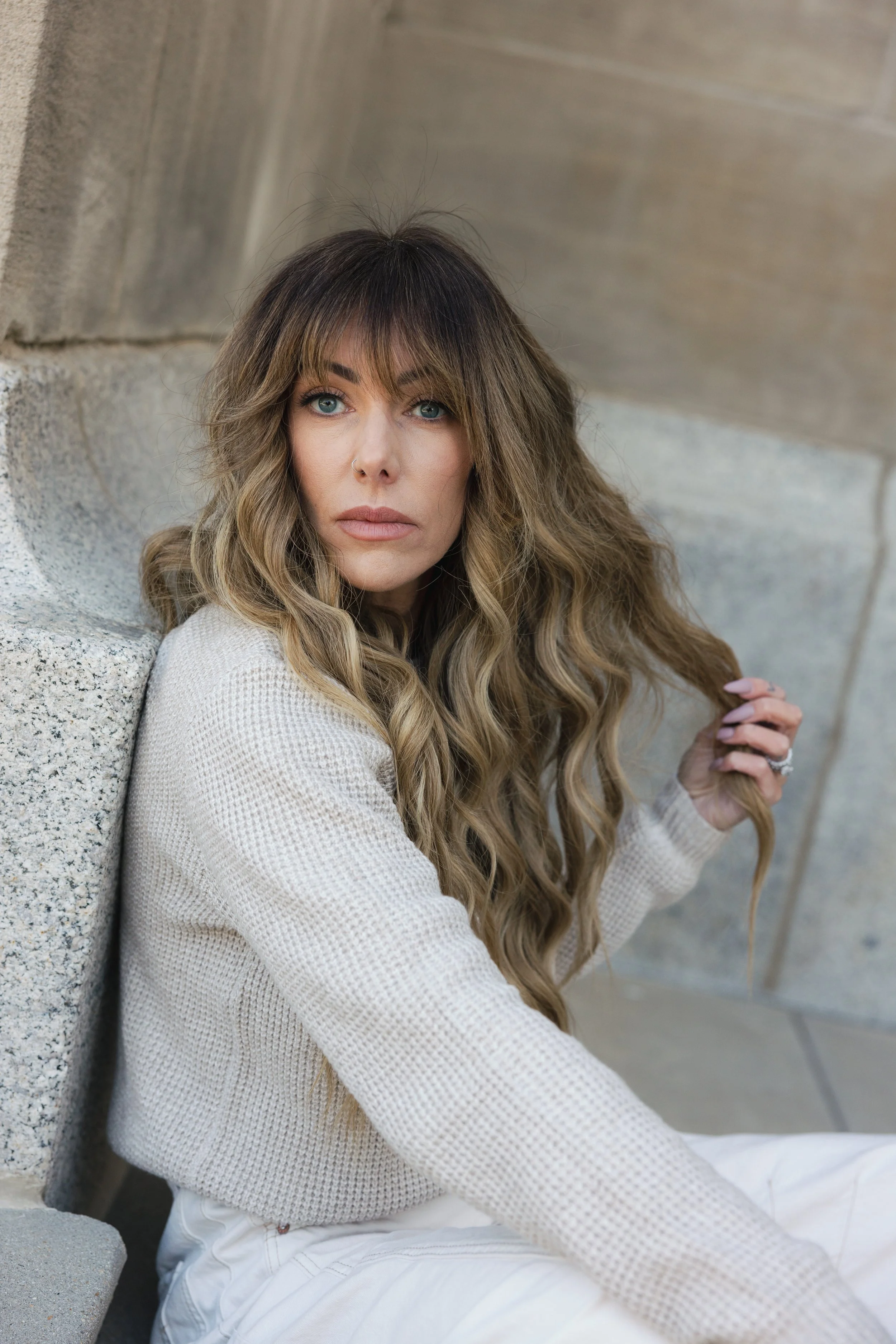 A woman with long, wavy blonde hair and blue eyes, sitting against a concrete wall, wearing a cream sweater and white pants, holding a strand of her hair, looking directly at the camera.