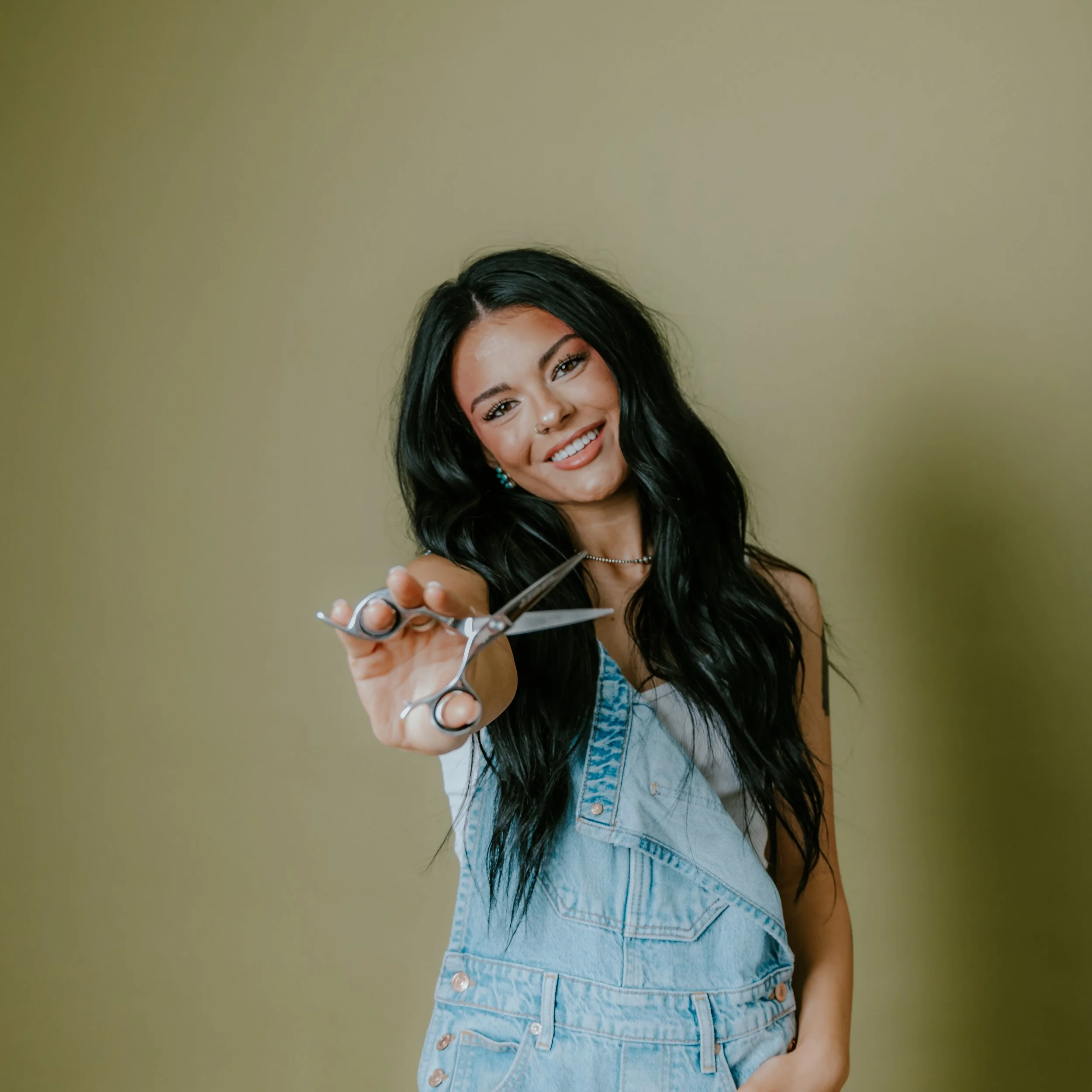 A young woman with long, dark, wavy hair smiling and holding a pair of scissors towards the camera.
