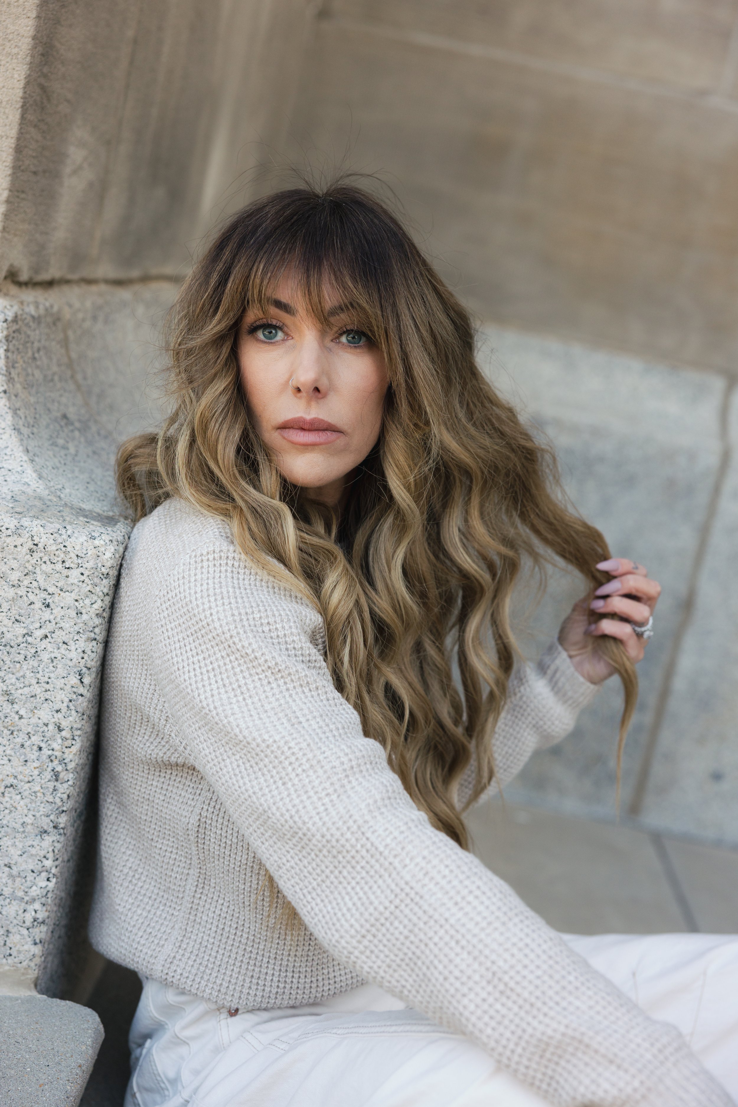 A woman with long wavy blonde hair and blue eyes, wearing a cream-colored sweater, sitting against a concrete wall and looking into the camera.