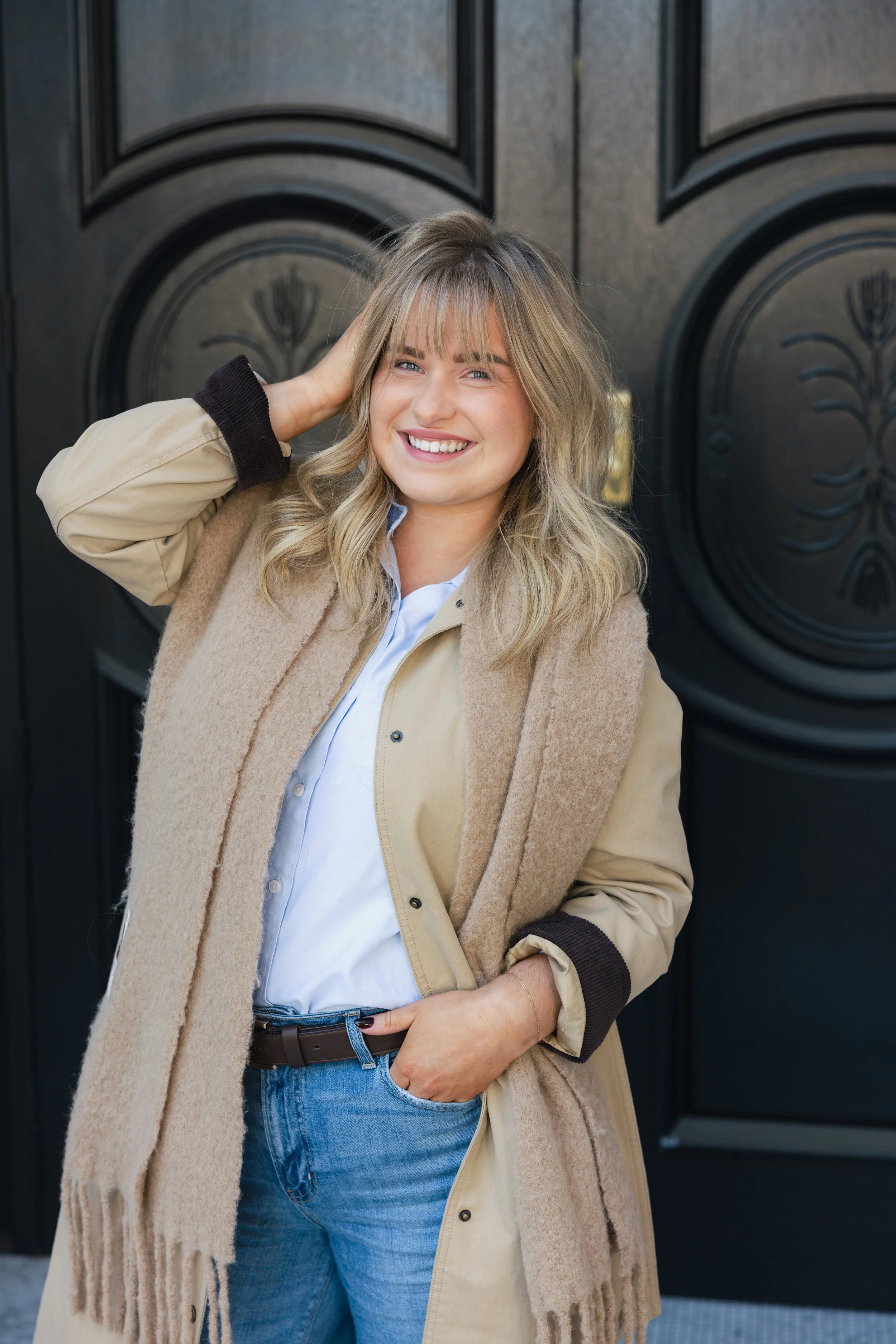 A smiling woman with blonde wavy hair, wearing a beige coat, white shirt, blue jeans, and a brown scarf, standing in front of a black door.