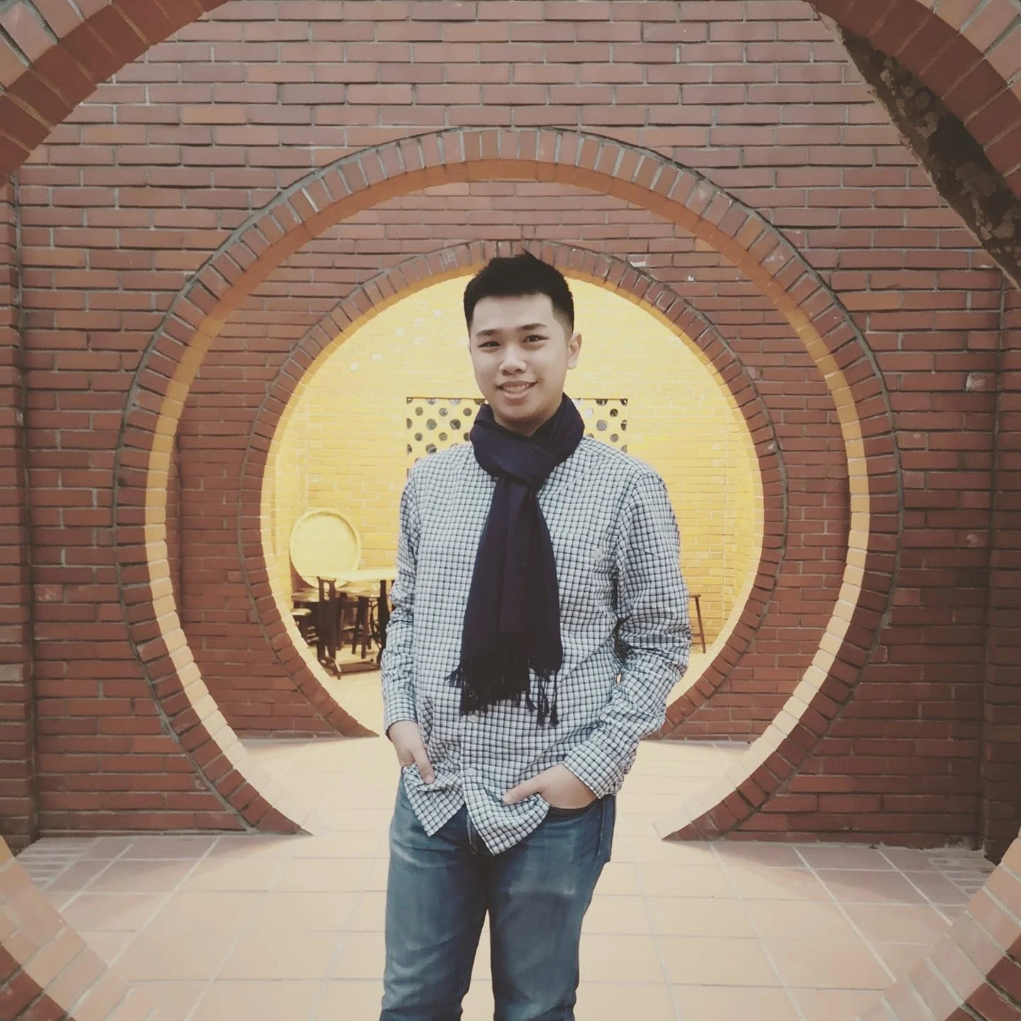 Young man standing in front of a decorative brick wall with circular openings, smiling and wearing a checked shirt and a dark scarf.