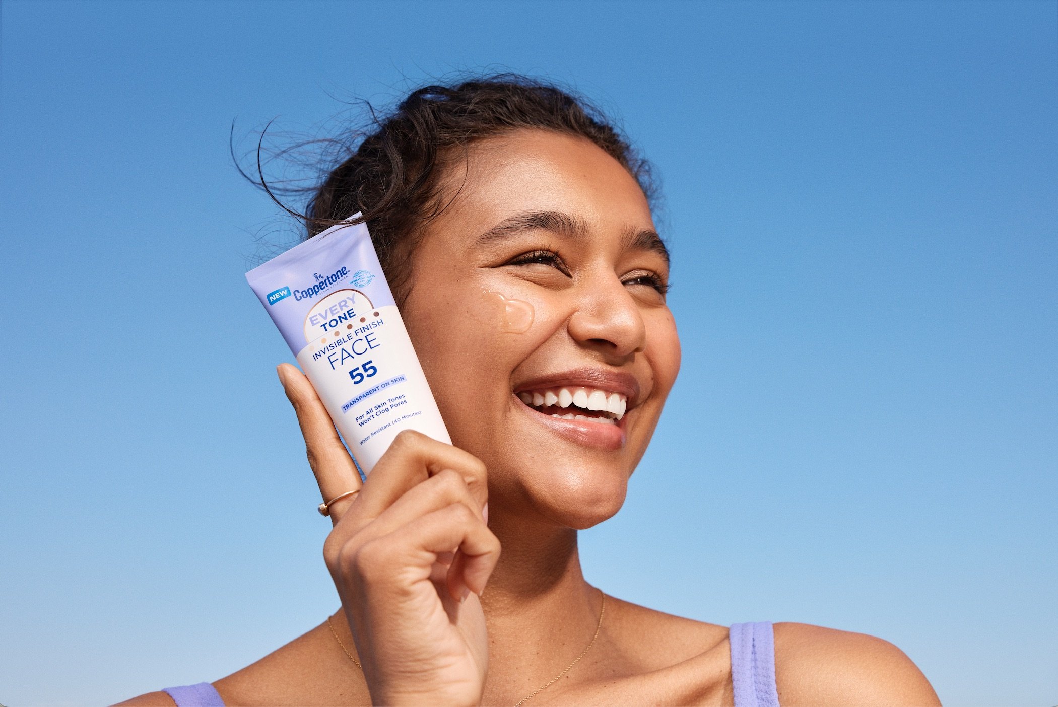 A smiling woman with dark curly hair outdoors against a clear blue sky, holding a tube of Coppertone sunscreen near her face.