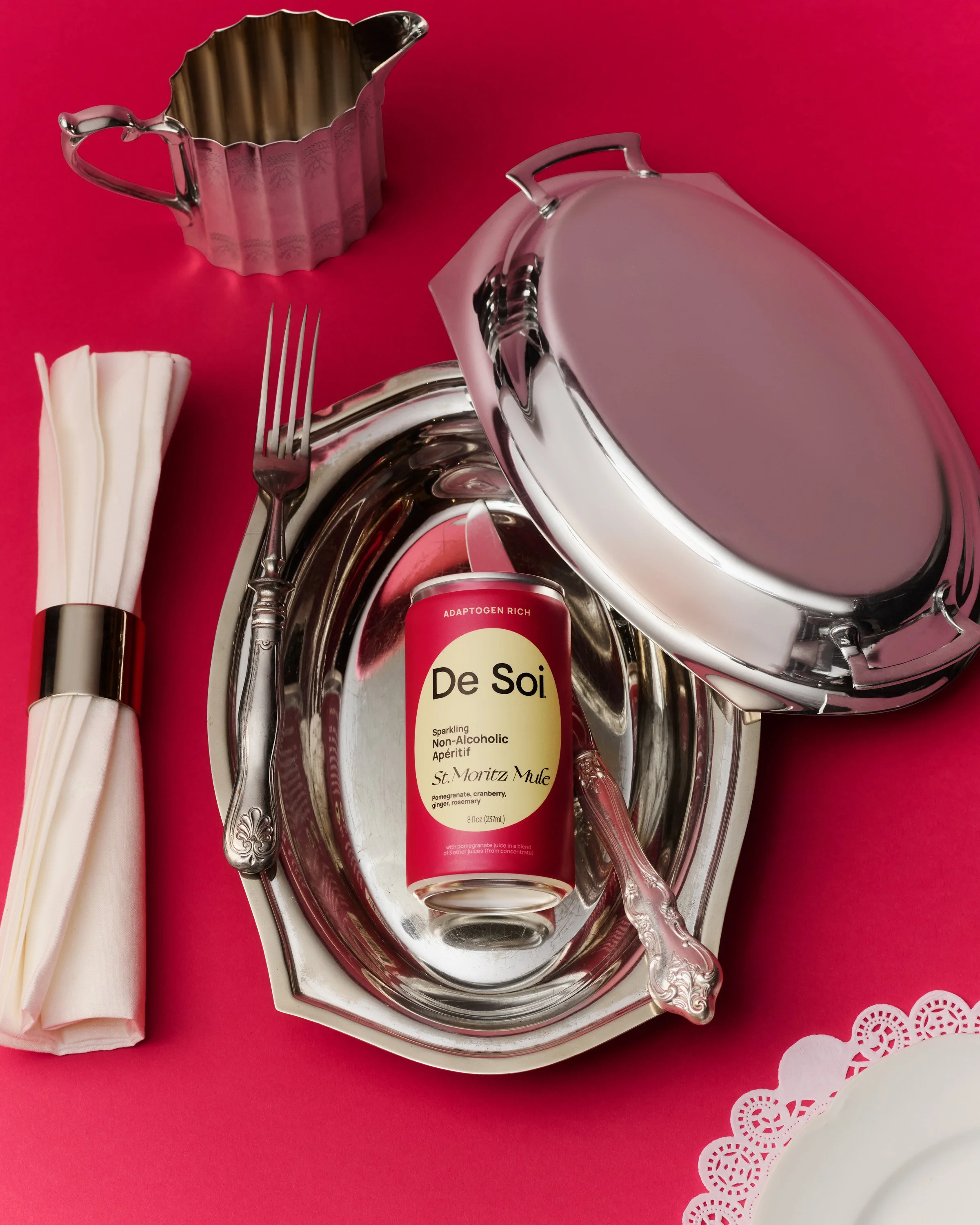 A table setting with a pink background, featuring a silver tray, a silver spoon, a fork, a wrapped napkin, a silver oval dish, a pink canned beverage labeled 'De Soi,' and a small silver cup.