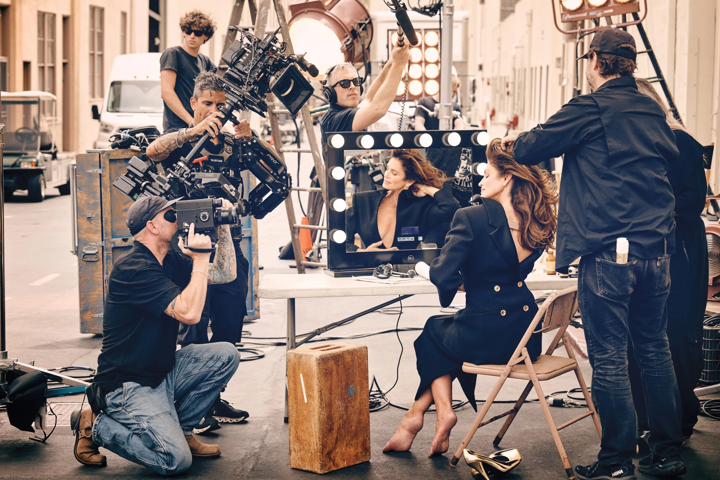 Behind the scenes of a photo shoot with two women sitting at a makeup station, surrounded by a crew of photographers and assistants in an outdoor urban setting.