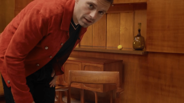 Man leaning over a dining table with wooden chairs in a wood-paneled room, with a yellow tennis ball and a decorative bottle on the shelf in the background.