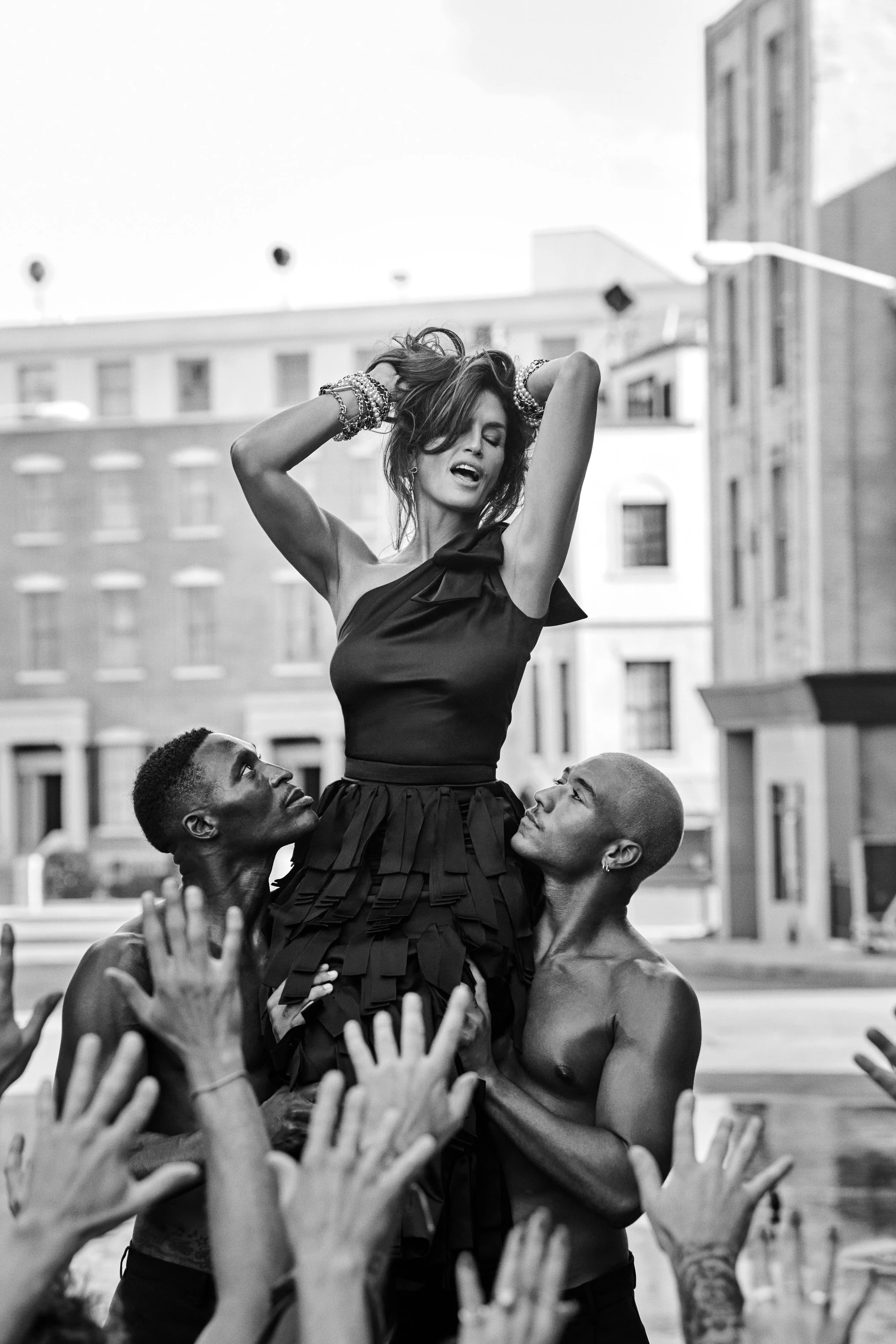 Black and white photograph of a woman with arms raised and eyes closed, being lifted by two shirtless men with hands reaching up towards her, set against a city street background.