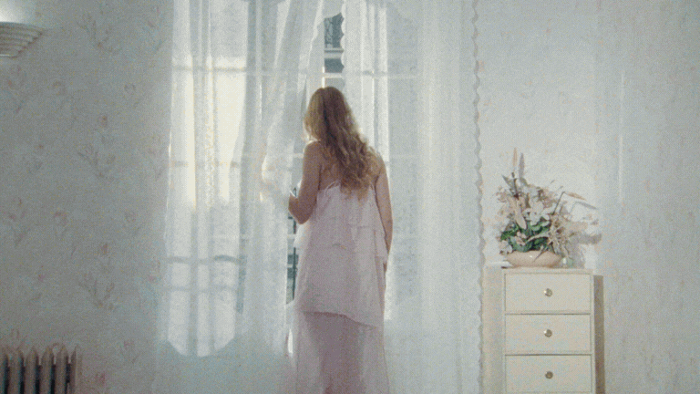 A woman with long blonde hair in a pink dress standing by a window with sheer curtains, looking outside in a softly lit room.