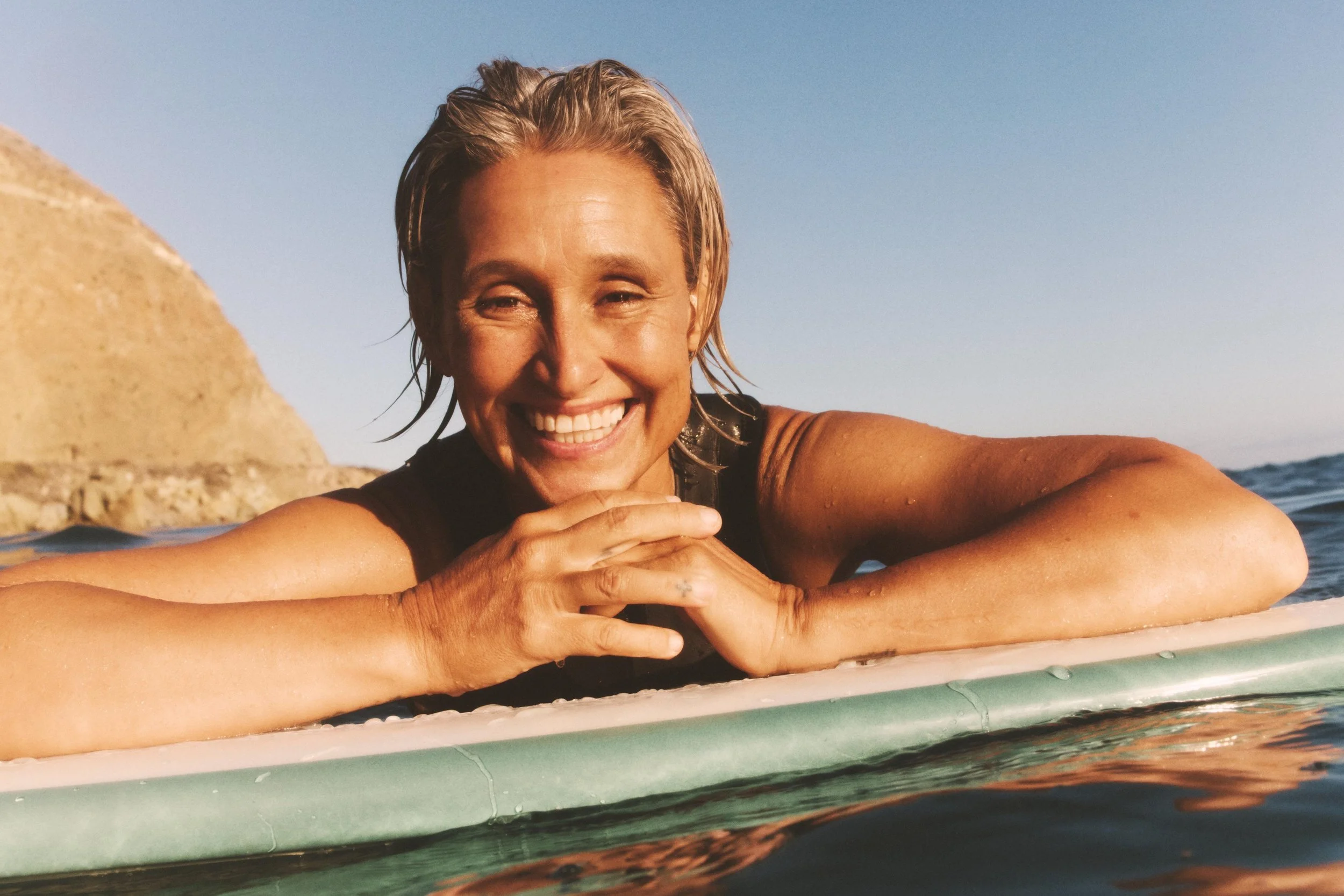 A woman with blonde hair smiling while lying on a surfboard in the water during sunset.