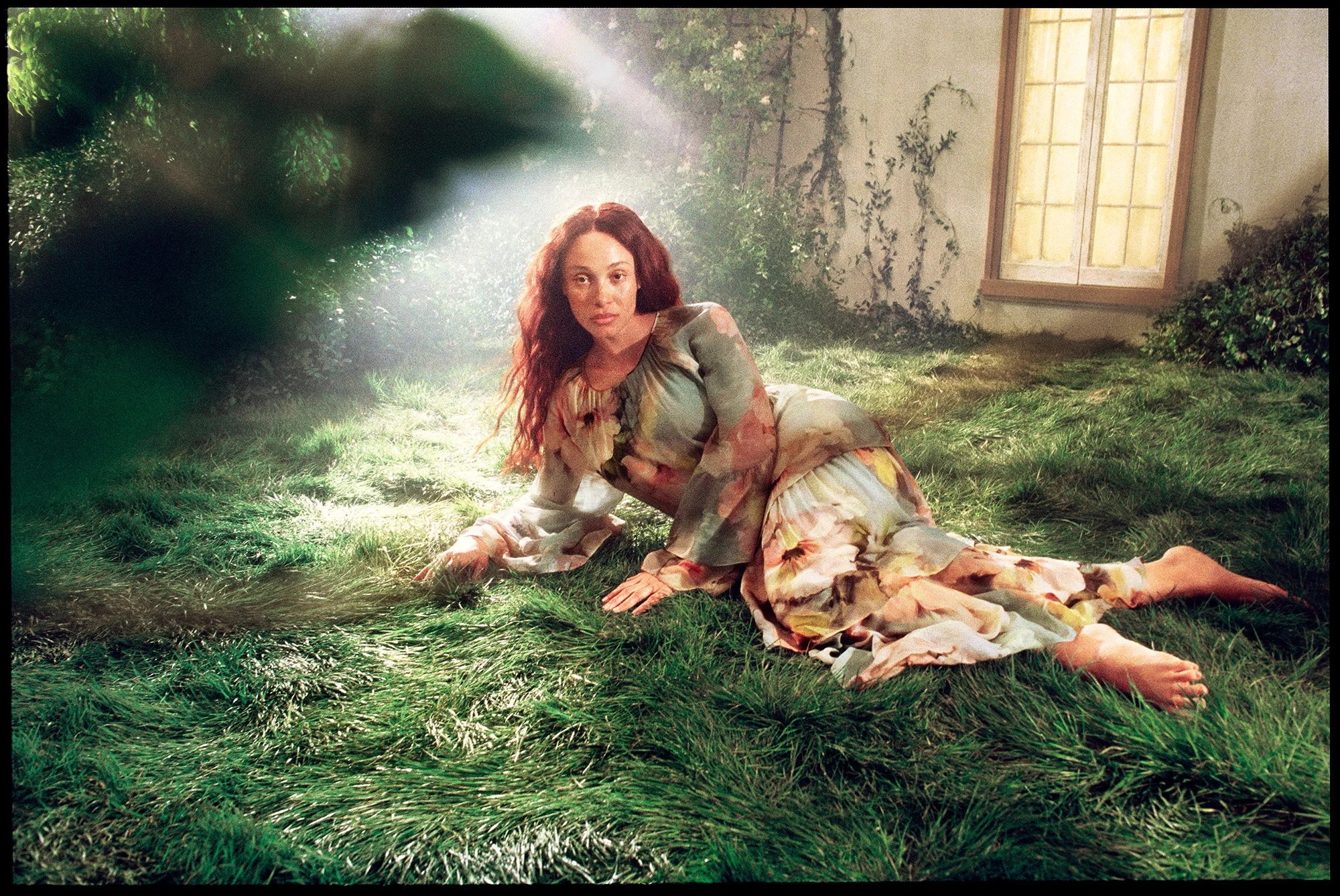 A woman with long, wavy red hair lying on grass in a sunlit room, wearing a flowing, floral-patterned dress, with an open window and ivy on the wall in the background.