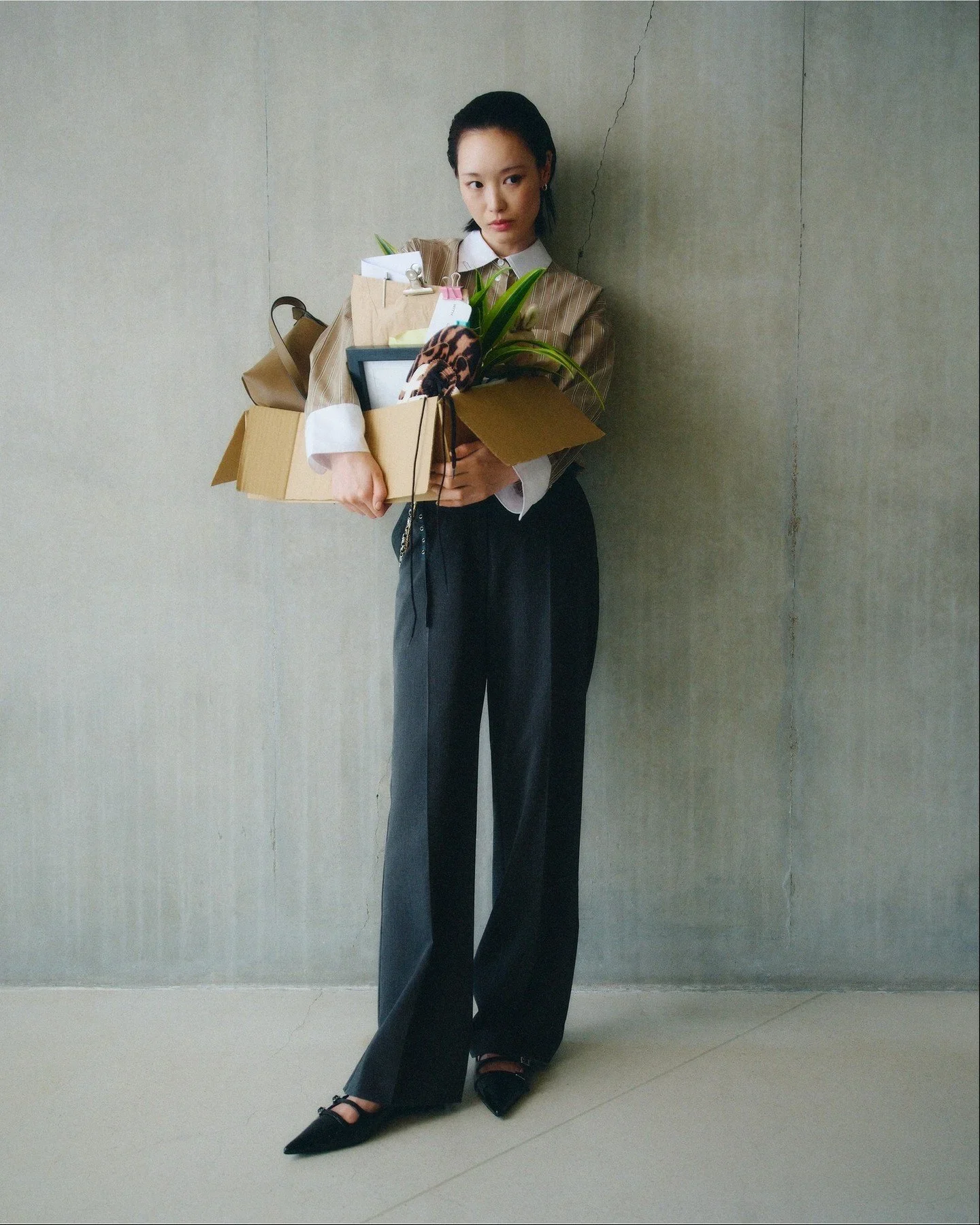 A woman dressed in business attire standing against a plain wall, holding a large box filled with office supplies, a potted plant, and a framed photo.
