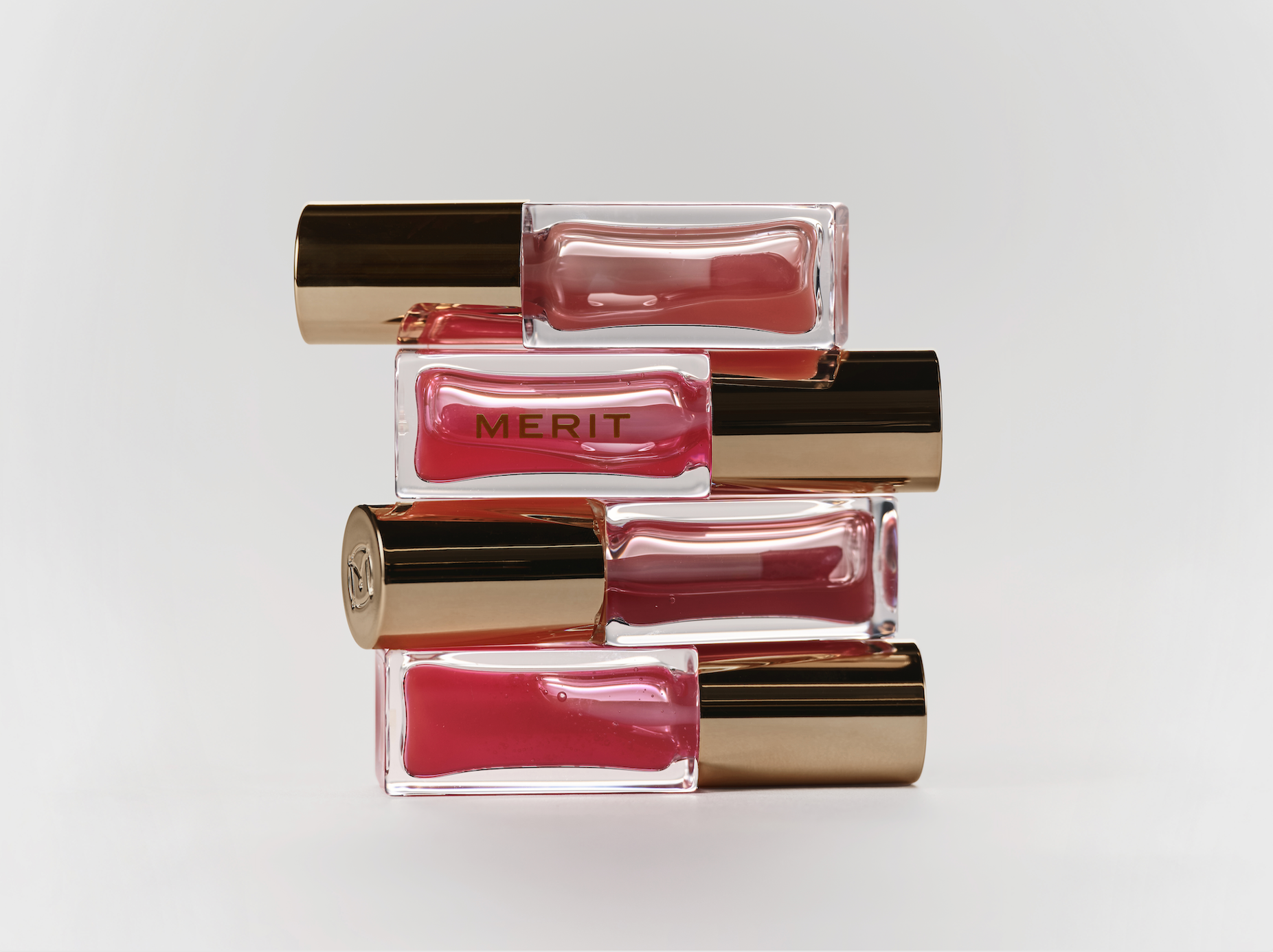 Stacked rectangular lip gloss bottles with gold caps, one labeled 'MERIT', with pinkish gloss inside, on a white background.