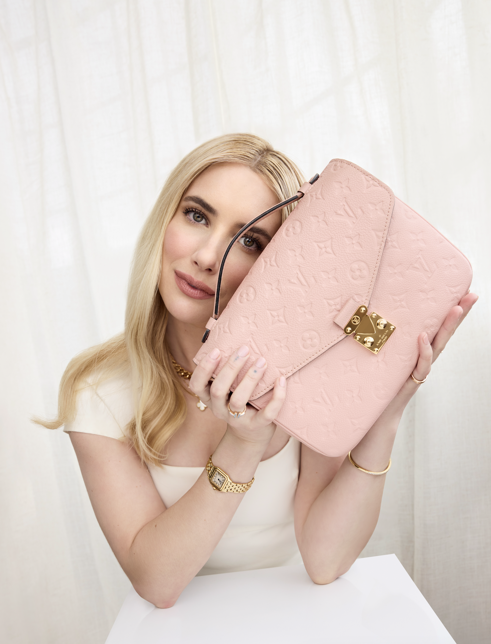 Woman with blond hair holding a pink designer handbag with a gold lock against a light-colored curtain background.