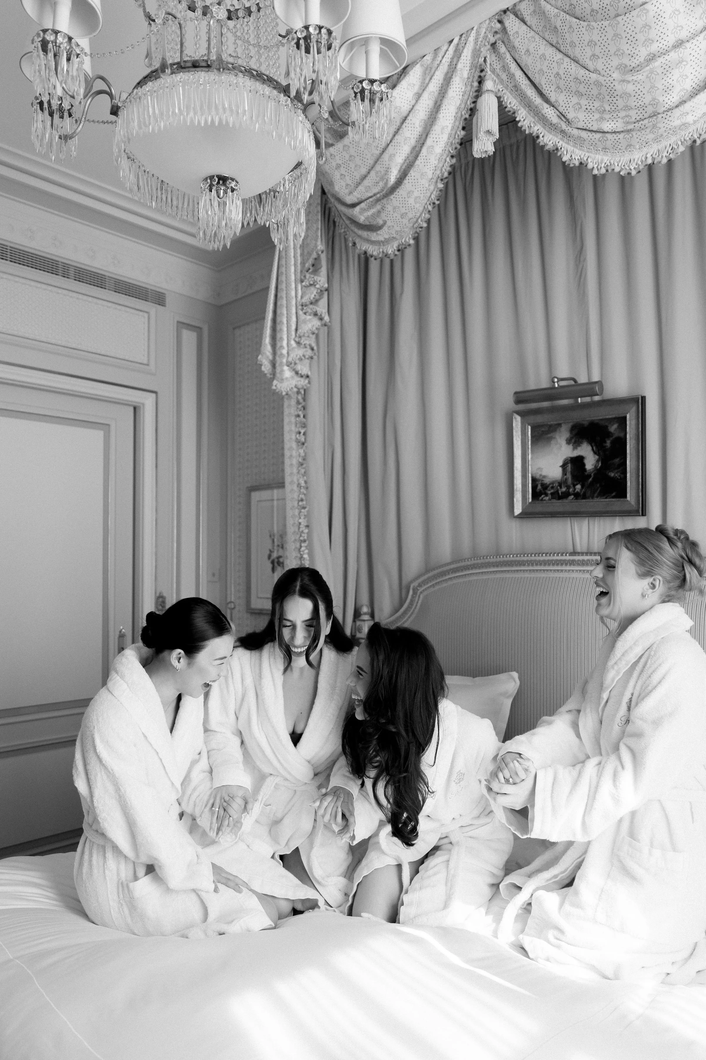 Four women in bathrobes sitting on a bed, laughing and holding hands, in an elegant hotel room with a chandelier and draped curtains.