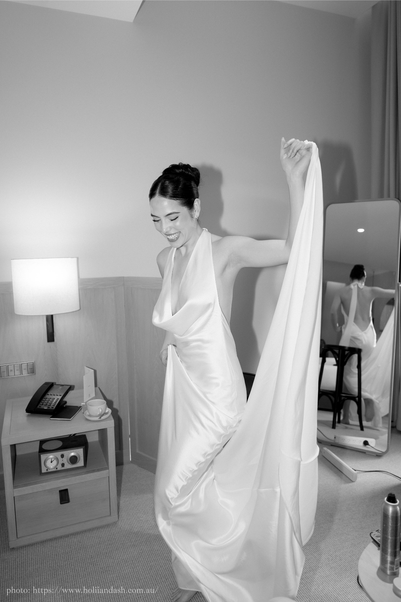 A woman in a satin dress is smiling and lifting a part of her dress in a hotel room, with her reflection visible in a mirror behind her.