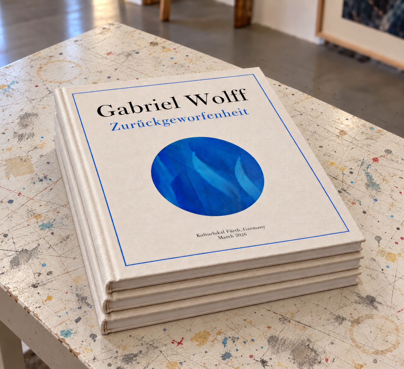 Stack of three cloth-bound copies of the exhibition catalogue Gabriel Wolff: Zurückgeworfenheit, with a blue circular image on the cover, on a paint-spattered worktable.