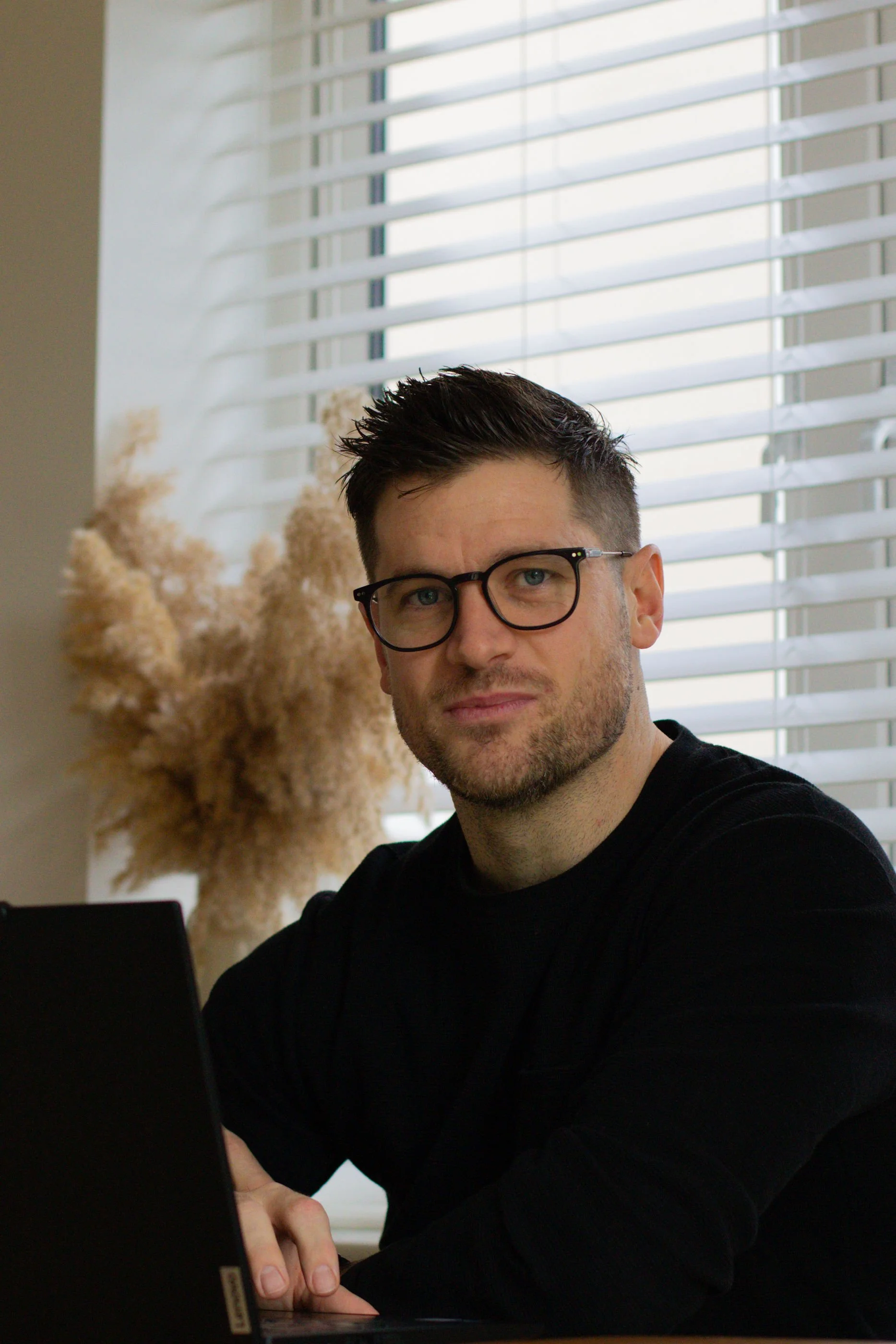 A man with dark hair, glasses, and a beard, sitting in front of a laptop with white blinds behind him.