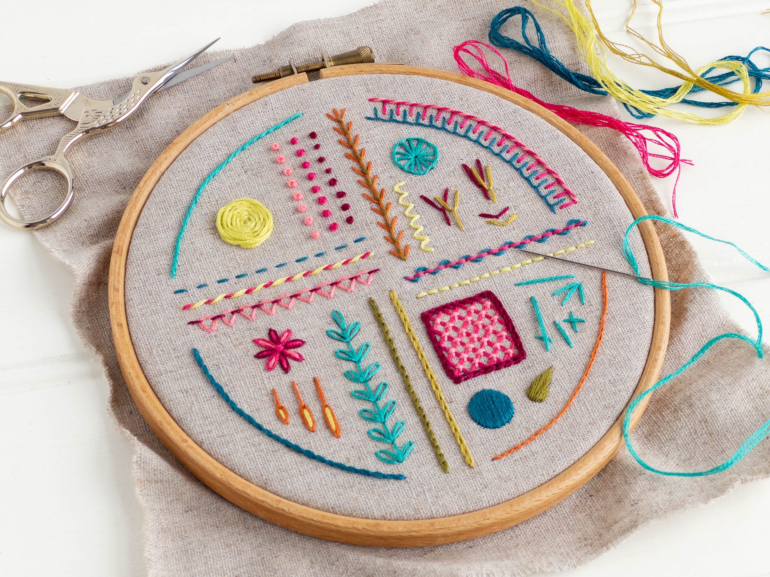 A hand-embroidered sampler worked in brightly coloured thread on natural linen is in a hoop, resting on a white workbench.