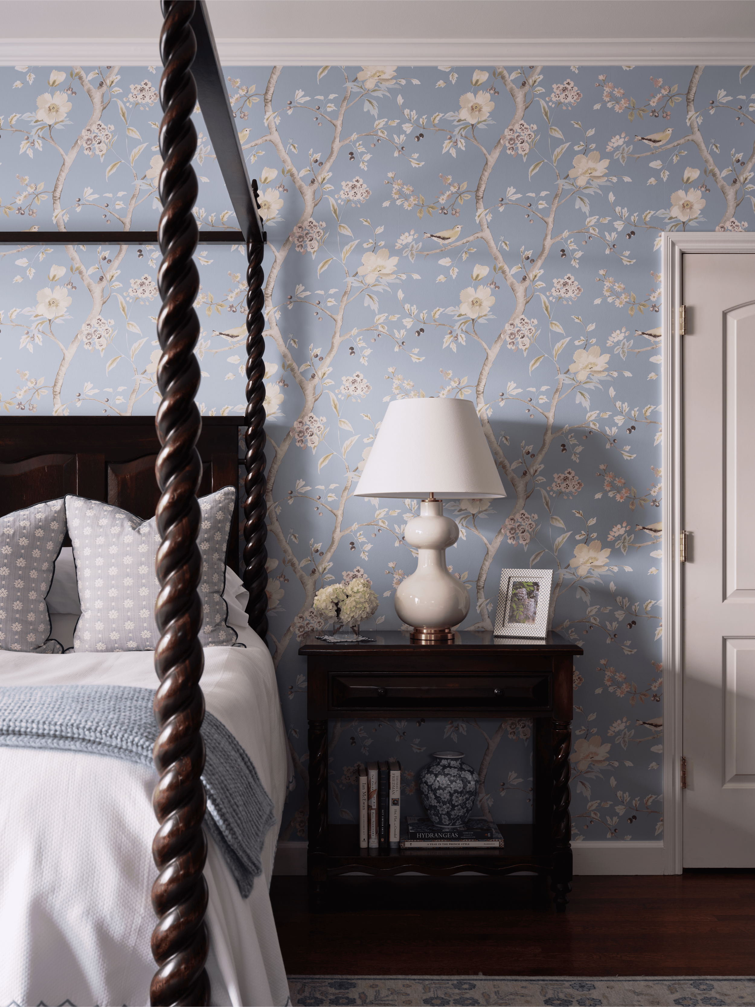 Kentfield Project bedroom designed by Arntz Interiors, featuring blue botanical wallpaper, a dark wood nightstand, white lamp, and canopy bedframe with spiral detail.