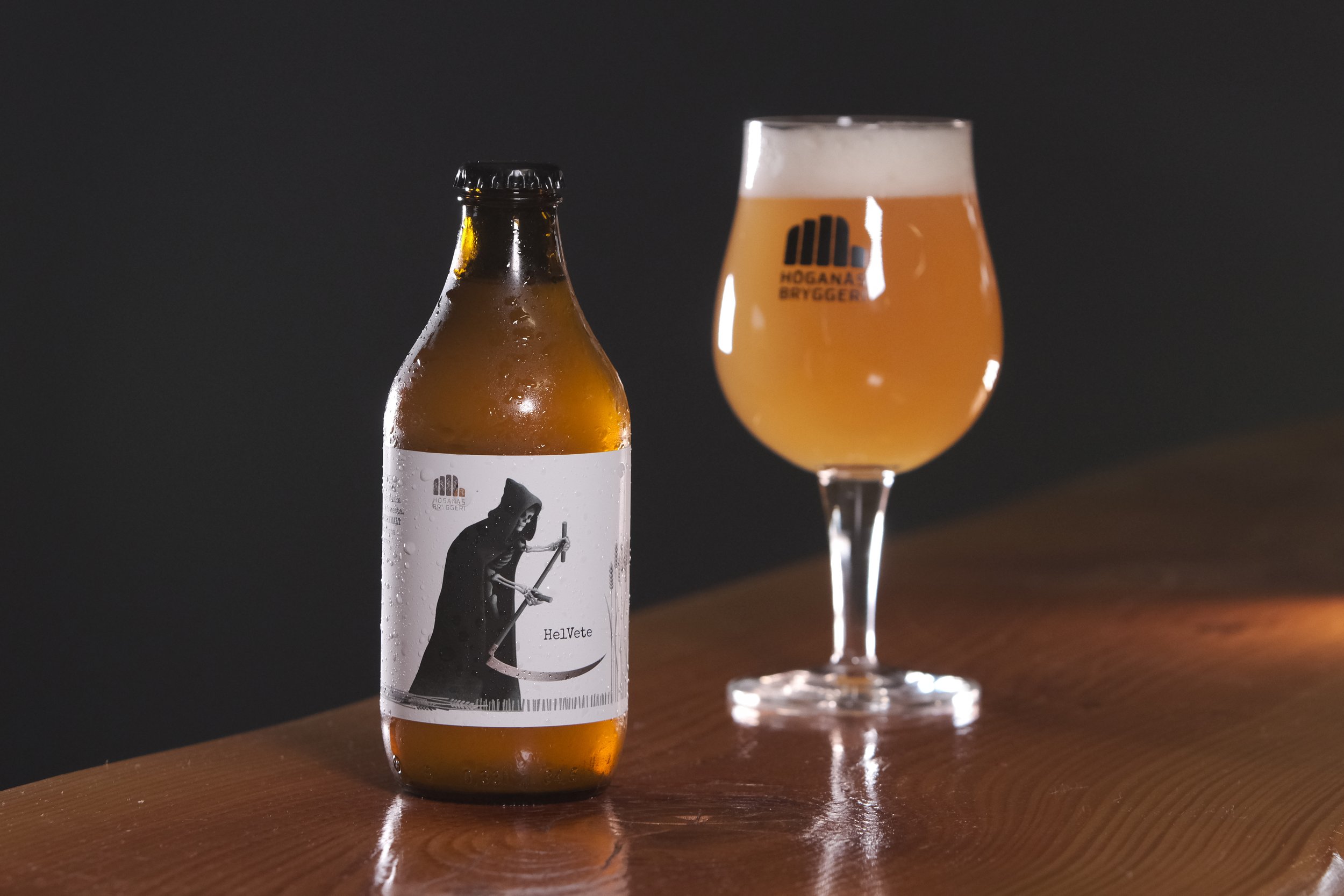 En ölflaska med etikett som visar en person i svart under en svart bakgrund, tillsammans med en öl i ett glas med ölens märke Högana Bryggeri. Flaskan står på ett träbord.