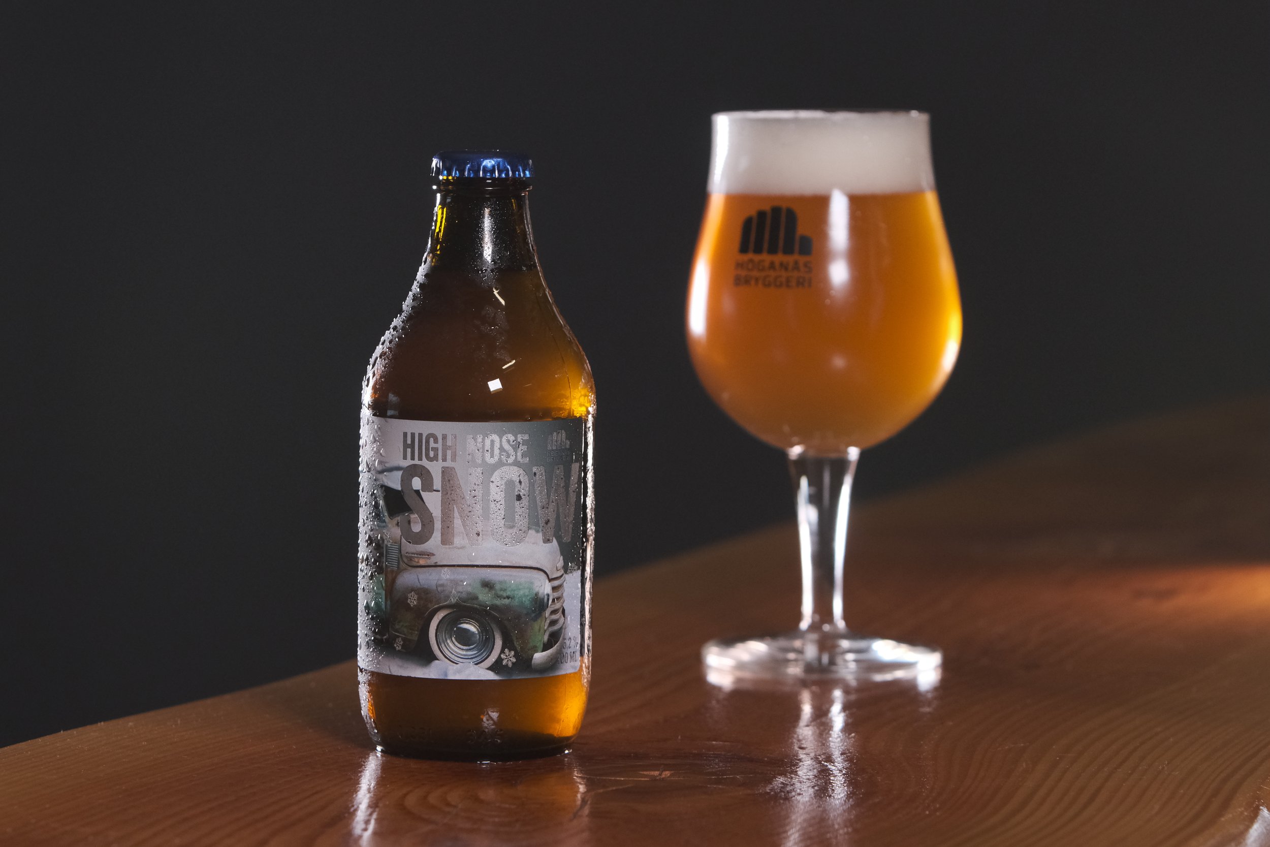 En burk med öl och ett ölglas på ett träbord, ölglaset har en logo som säger Hoganas Bryggeri, och burken har en etikett med texten High Nose Snow.