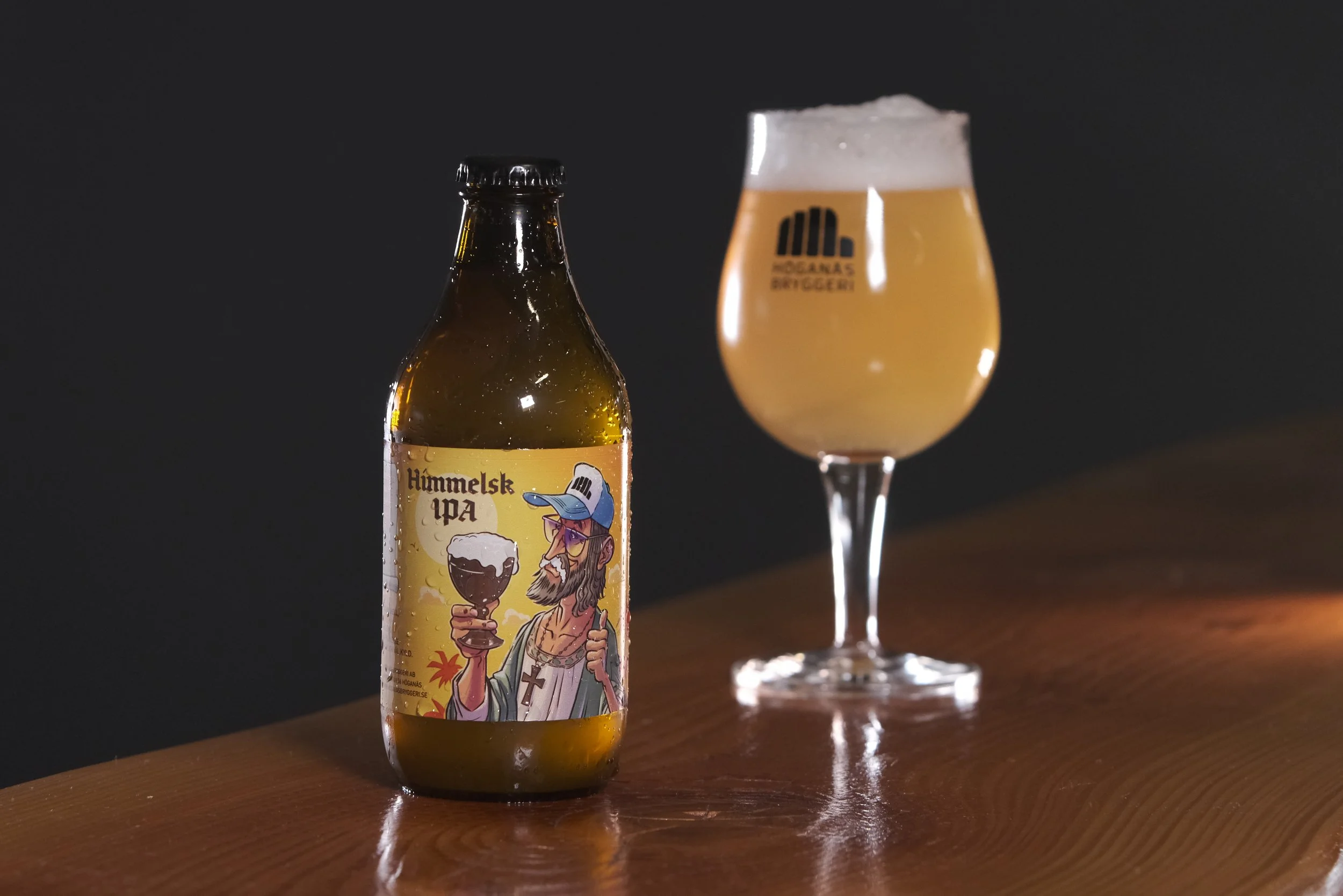 En ölflaska med en etikett som visar en tecknad man med lång hår och skägg, som håller i ett glasöl, samt ett glas fylld med öl bredvid flaskan på en träbord.
