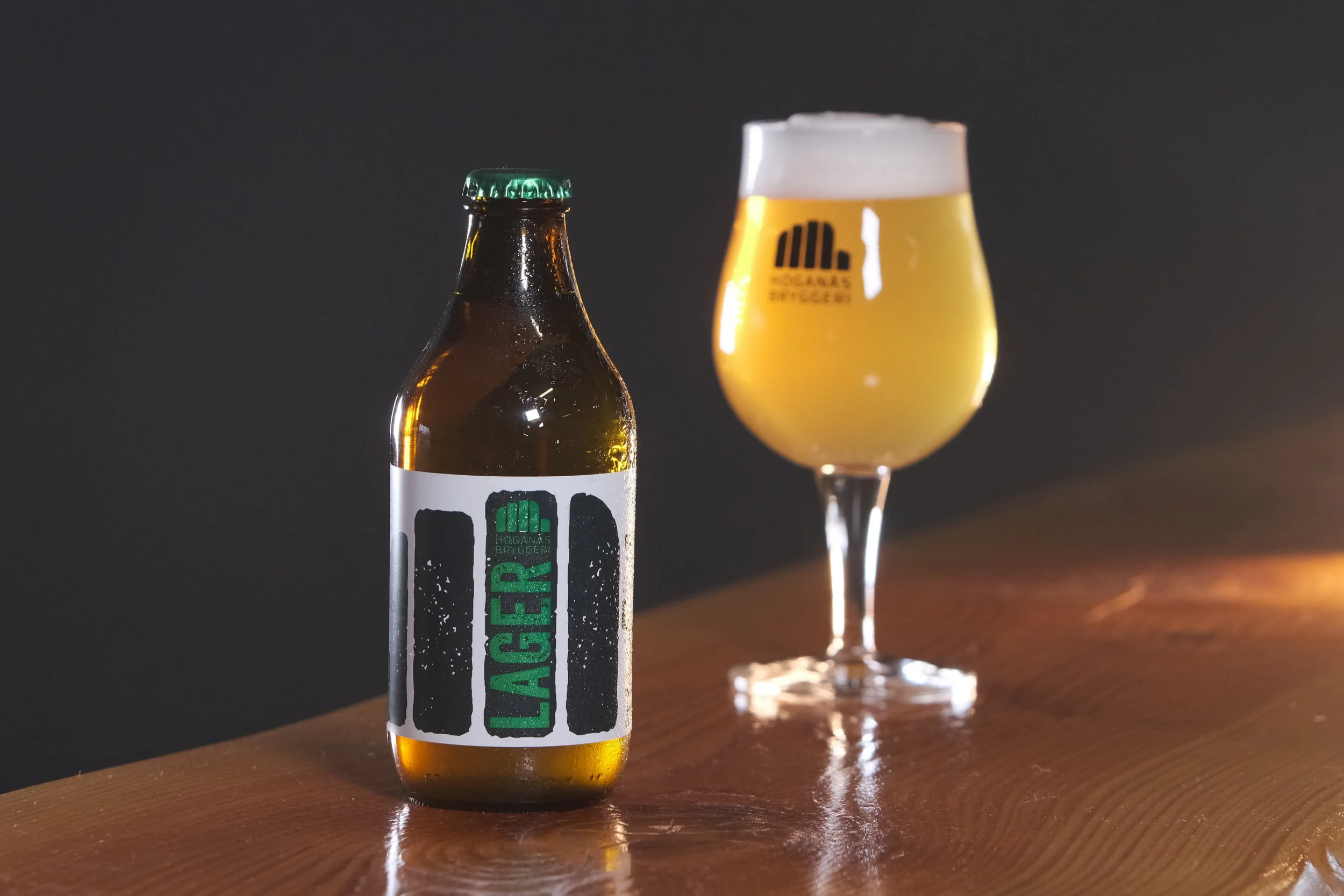 En ölflaska med en etikett som säger 'HOGAN'S BREWERY' och en ölglas med gyllene öl och ett lager av skum ovanpå, båda placerade på ett träbord mot en mörk bakgrund.