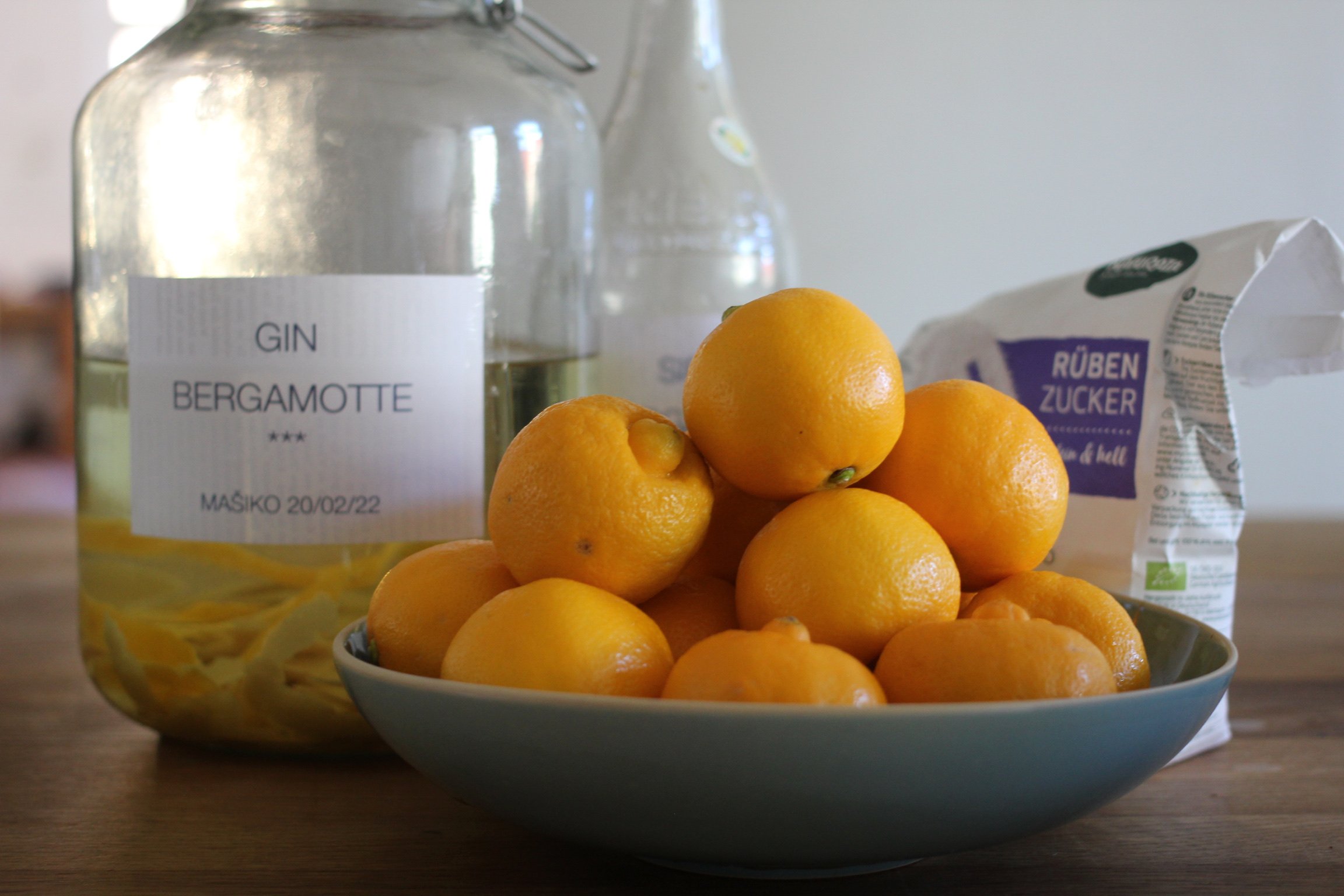 Ein Glas mit der Aufschrift "Gin Bergamotte", eine Schüssel voller Zitronen und eine Packung Rübenzucker stehen auf einem Tisch.