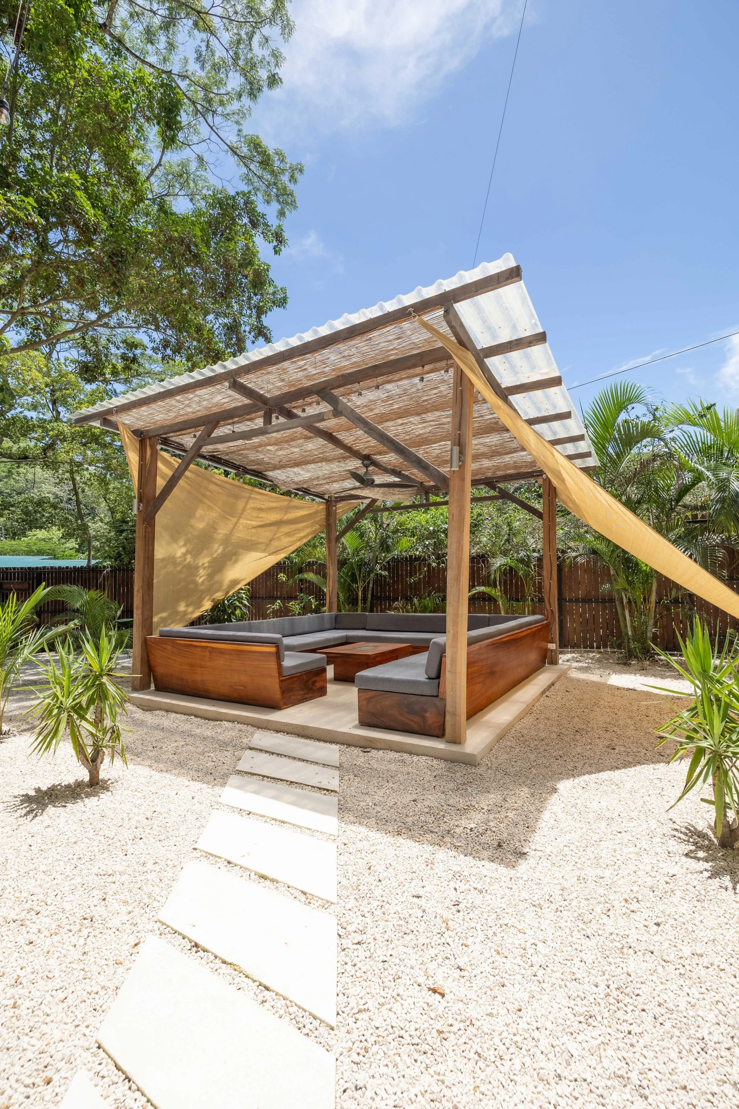 Outdoor patio area with wooden framed seating and gray cushions, a small wooden table, gravel ground, surrounded by tropical plants and trees, under a stone roof with shade sails, and a blue sky overhead.