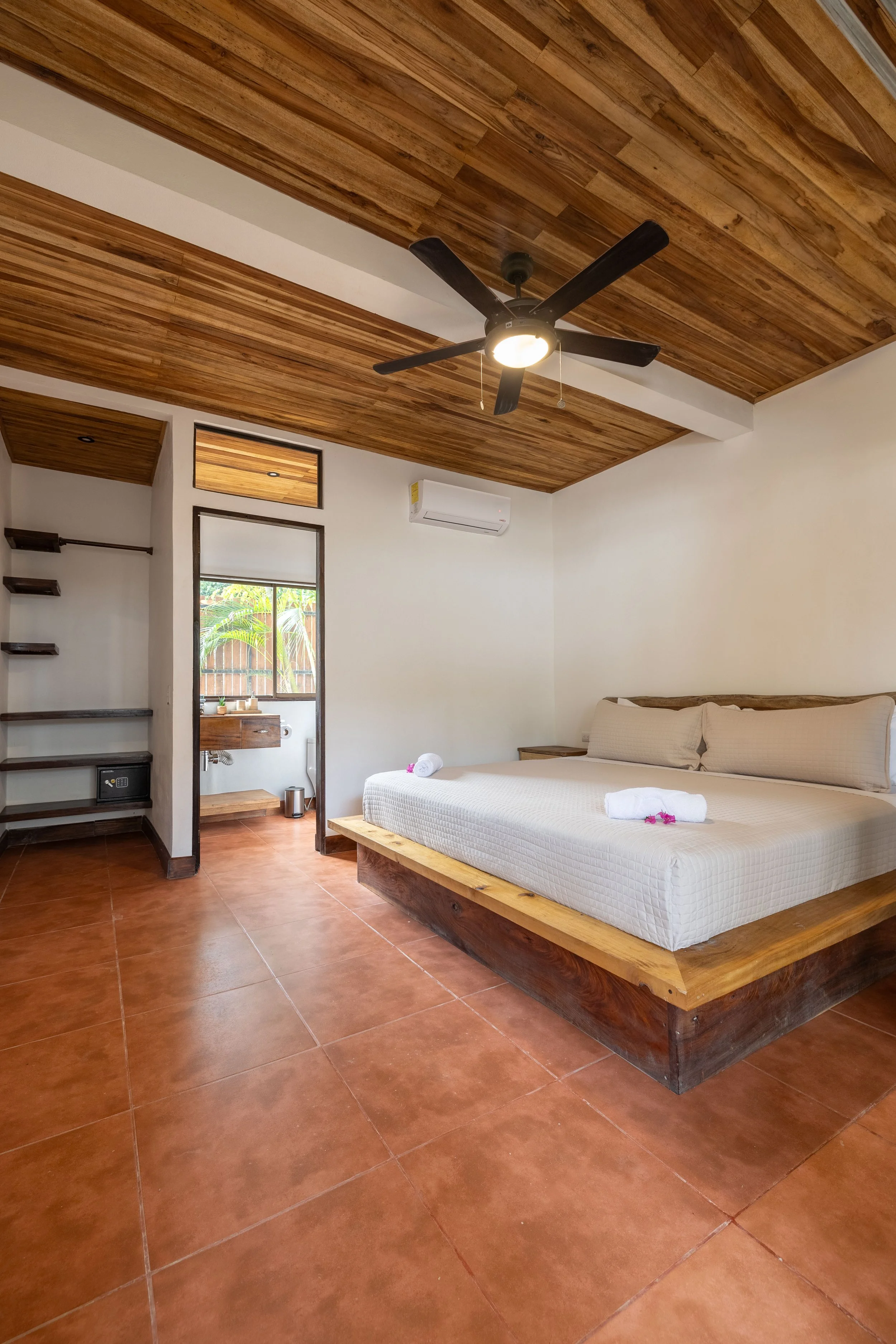 A modern bedroom with a wooden ceiling, a ceiling fan, white walls, terracotta tile flooring, a large bed with white linens, and a small bathroom visible through an open door.
