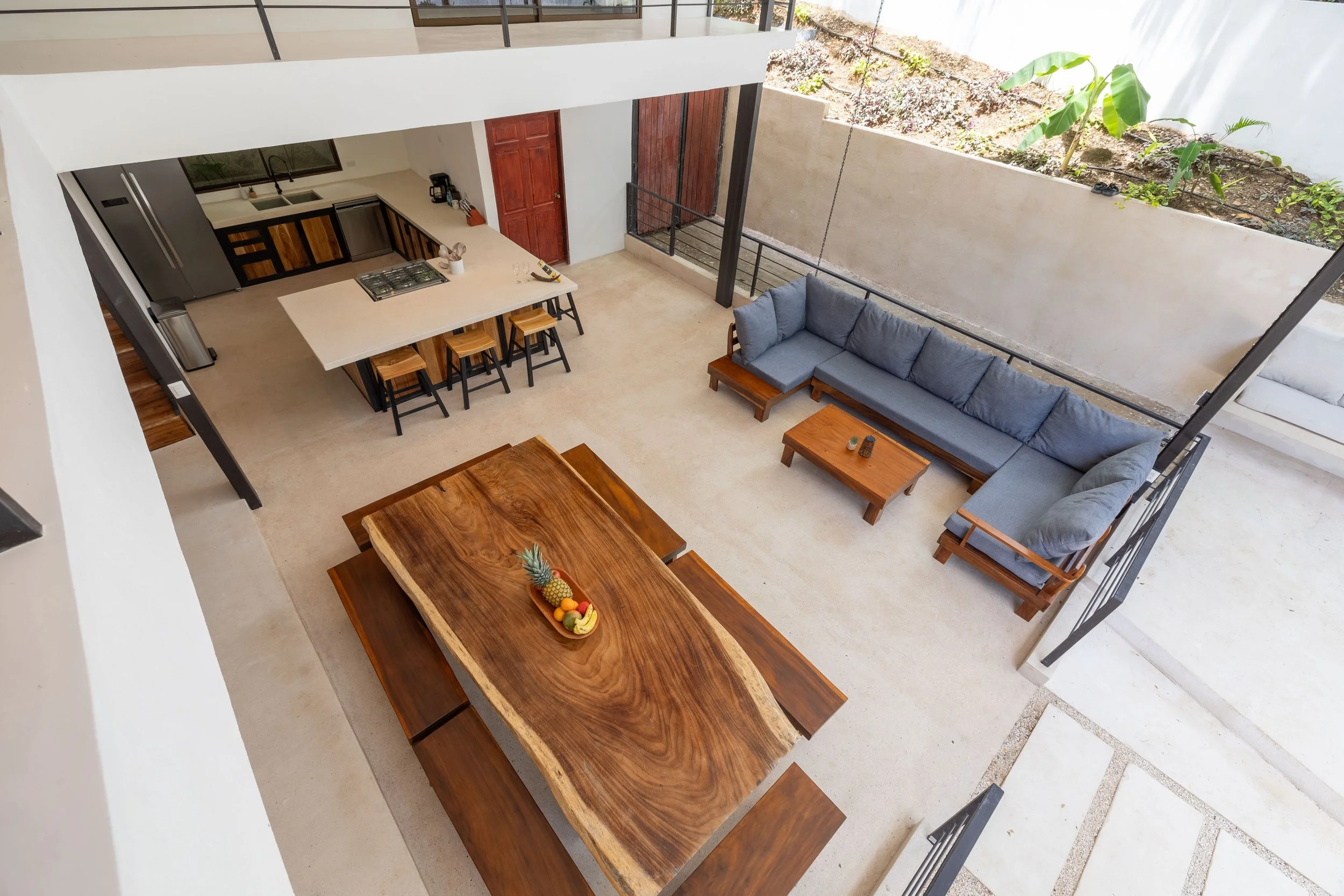 Open-concept living area with a kitchen, dining area, and lounge. The kitchen has black and wooden cabinets, a white countertop, and stainless steel appliances. The dining table is crafted from wood with matching benches, and the living space features a gray sectional sofa with a wooden coffee table. There is an outdoor patio with a garden area visible through a partial wall.
