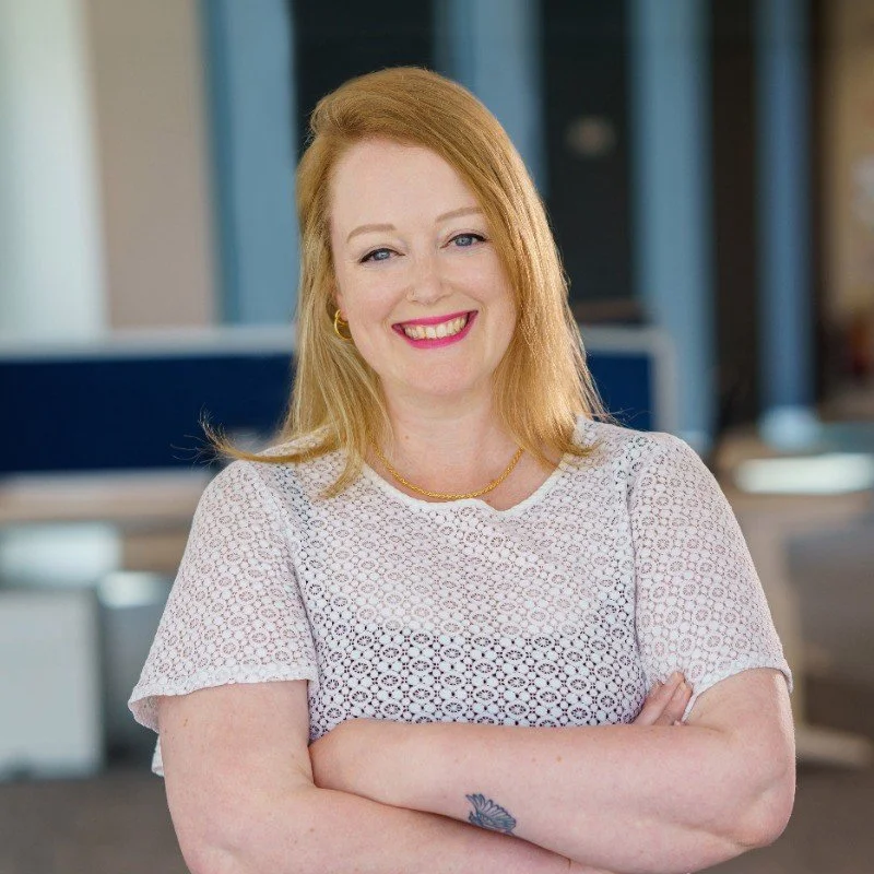 Professional woman with blonde hair, wearing a white crochet top and gold jewelry, smiling with crossed arms in an office environment.