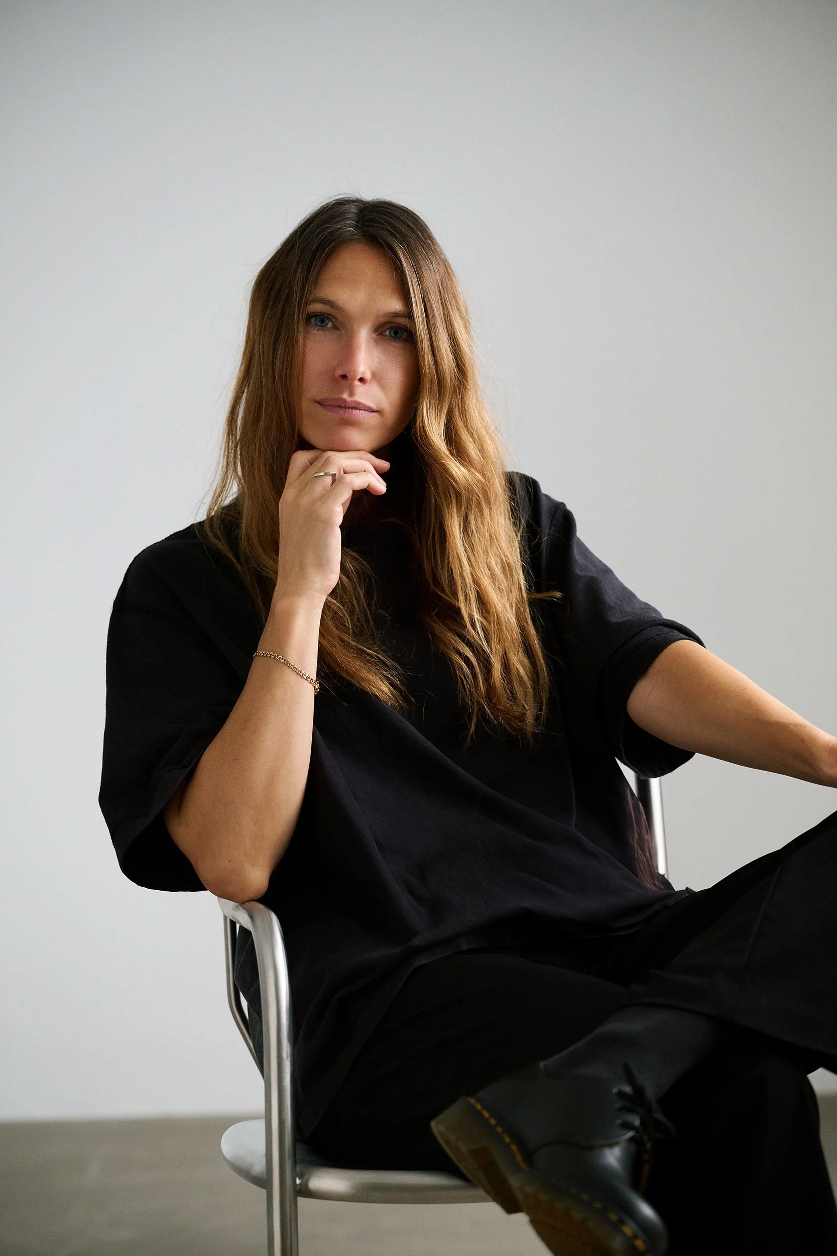 A woman sitting in a modern chair with a neutral expression, wearing a black t-shirt and pants, with long wavy brown hair and a silver bracelet, against a plain light gray background.