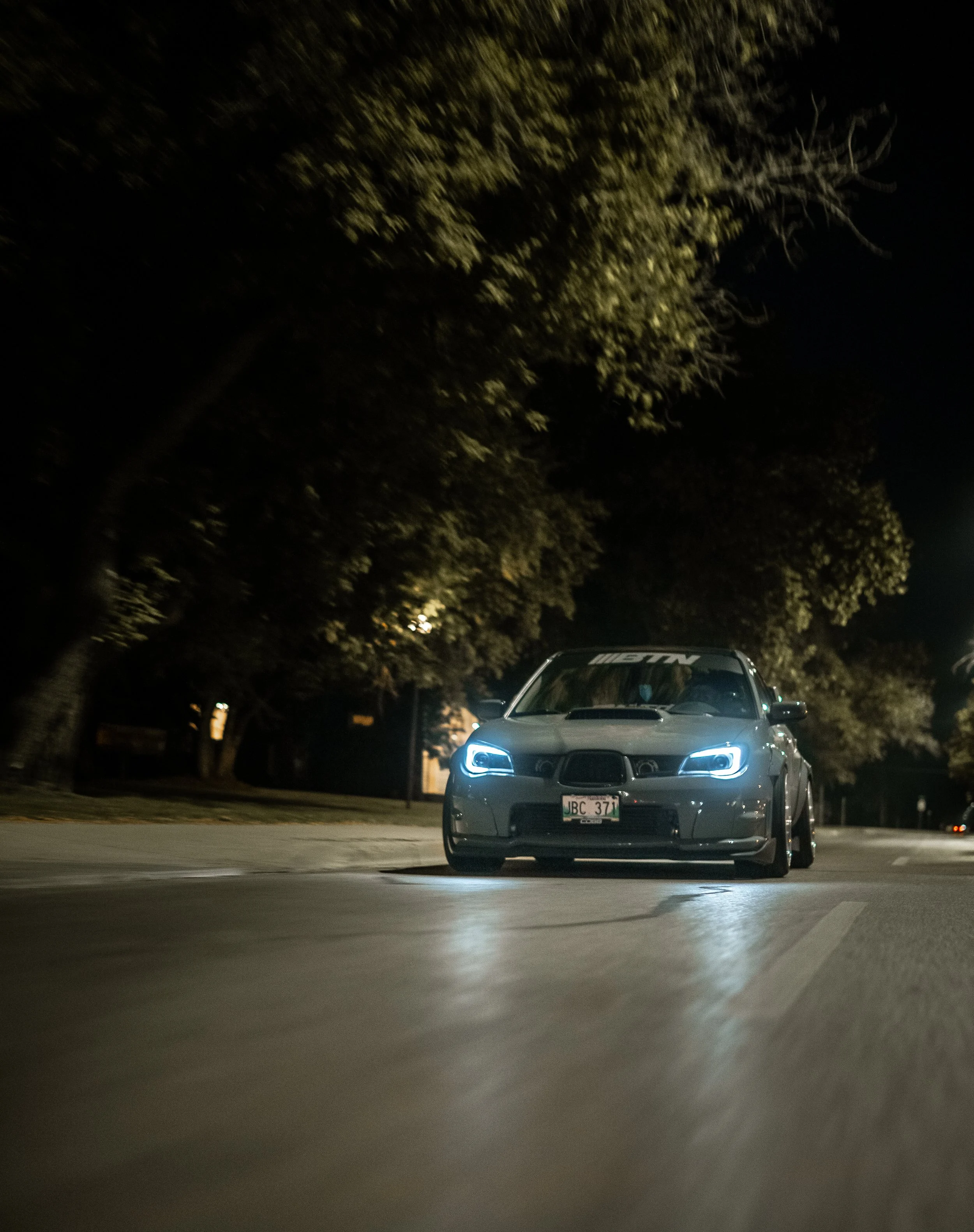 A dark nighttime photo of a grey sports car with bright headlights on an empty street, with trees in the background.