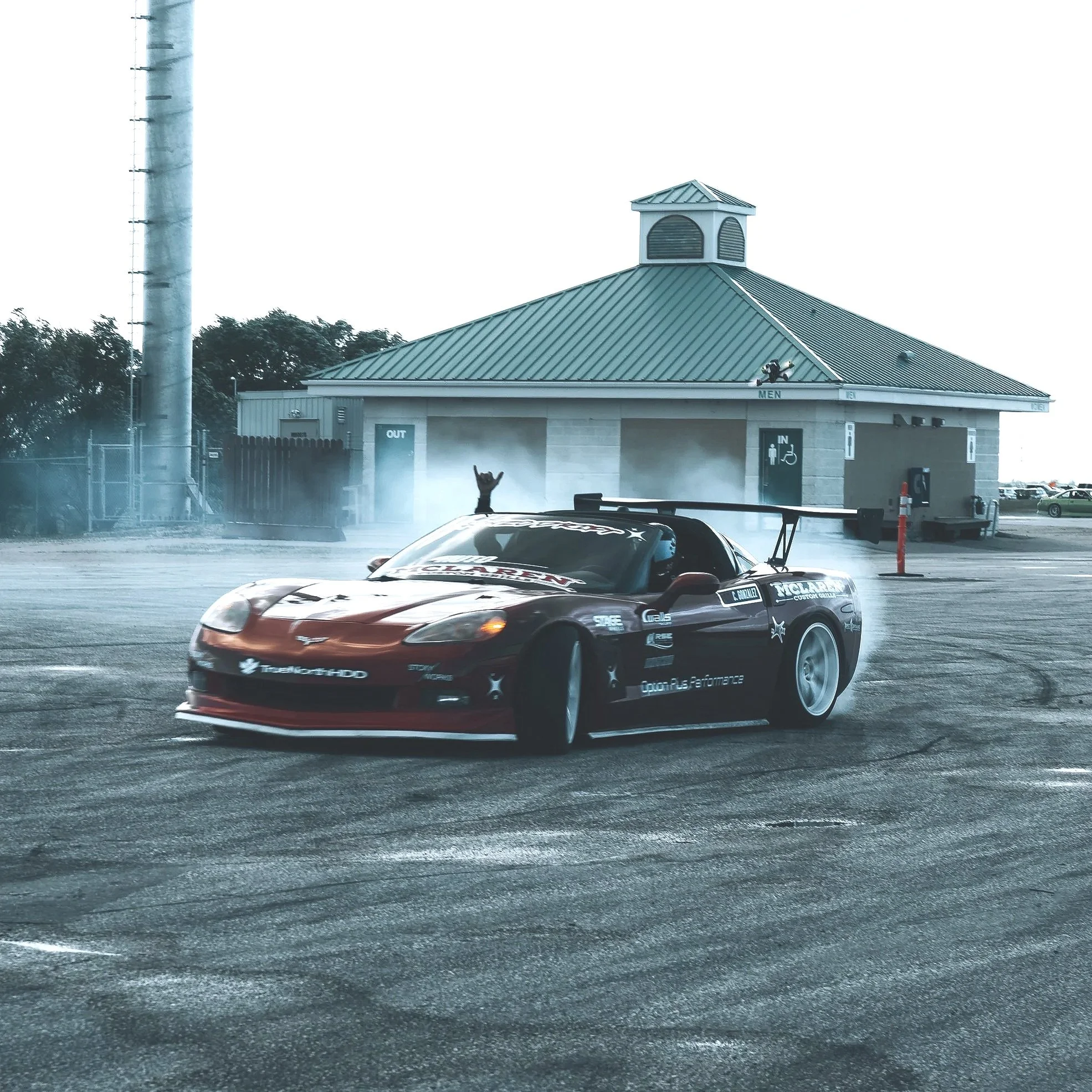 A race car performing a burnout in a parking lot with a building in the background and a person inside the car raising their hands.