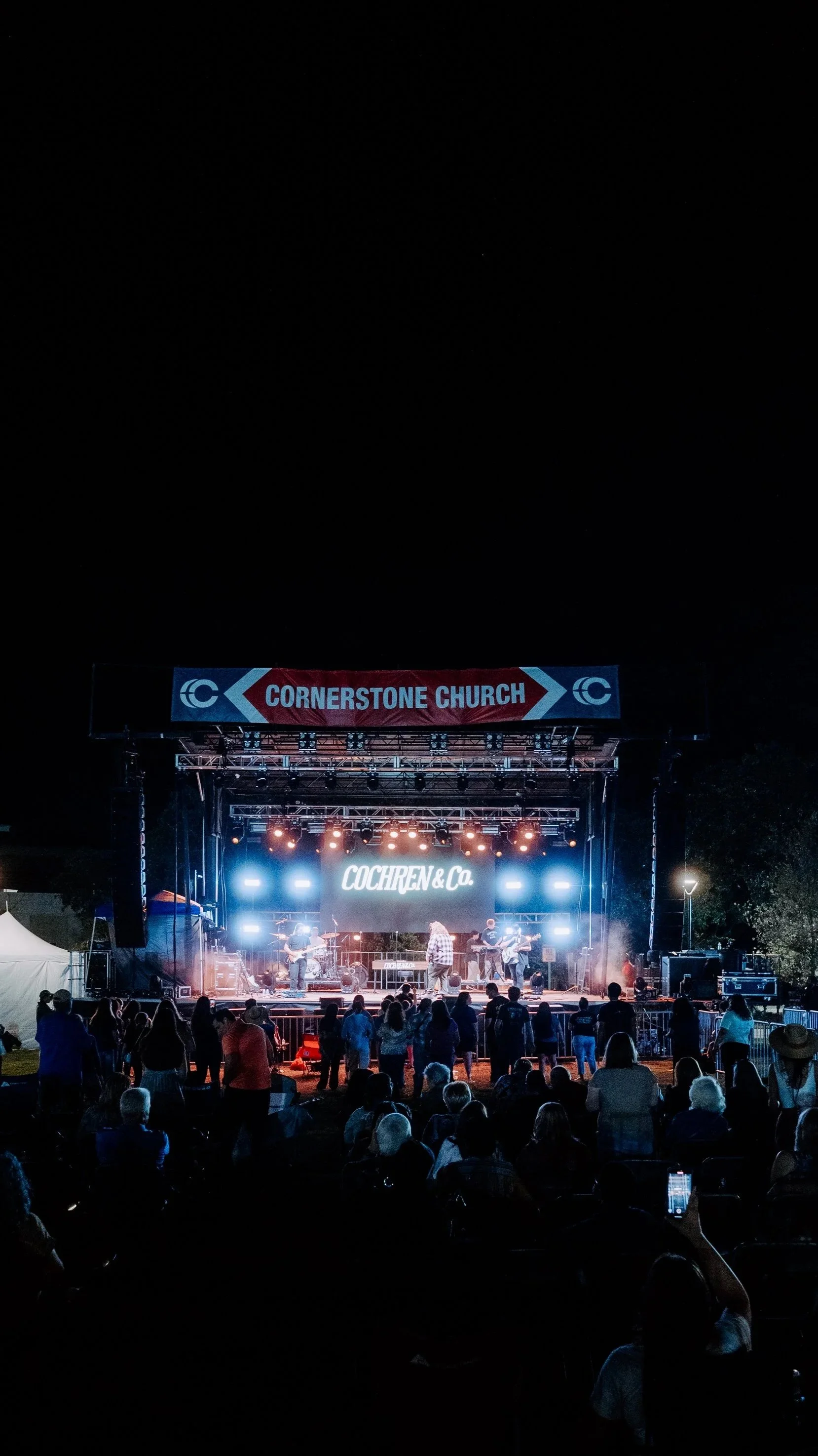 Nighttime outdoor concert with a band performing on stage, audience watching, and a sign reading 'Cornerstone Church' above the stage.