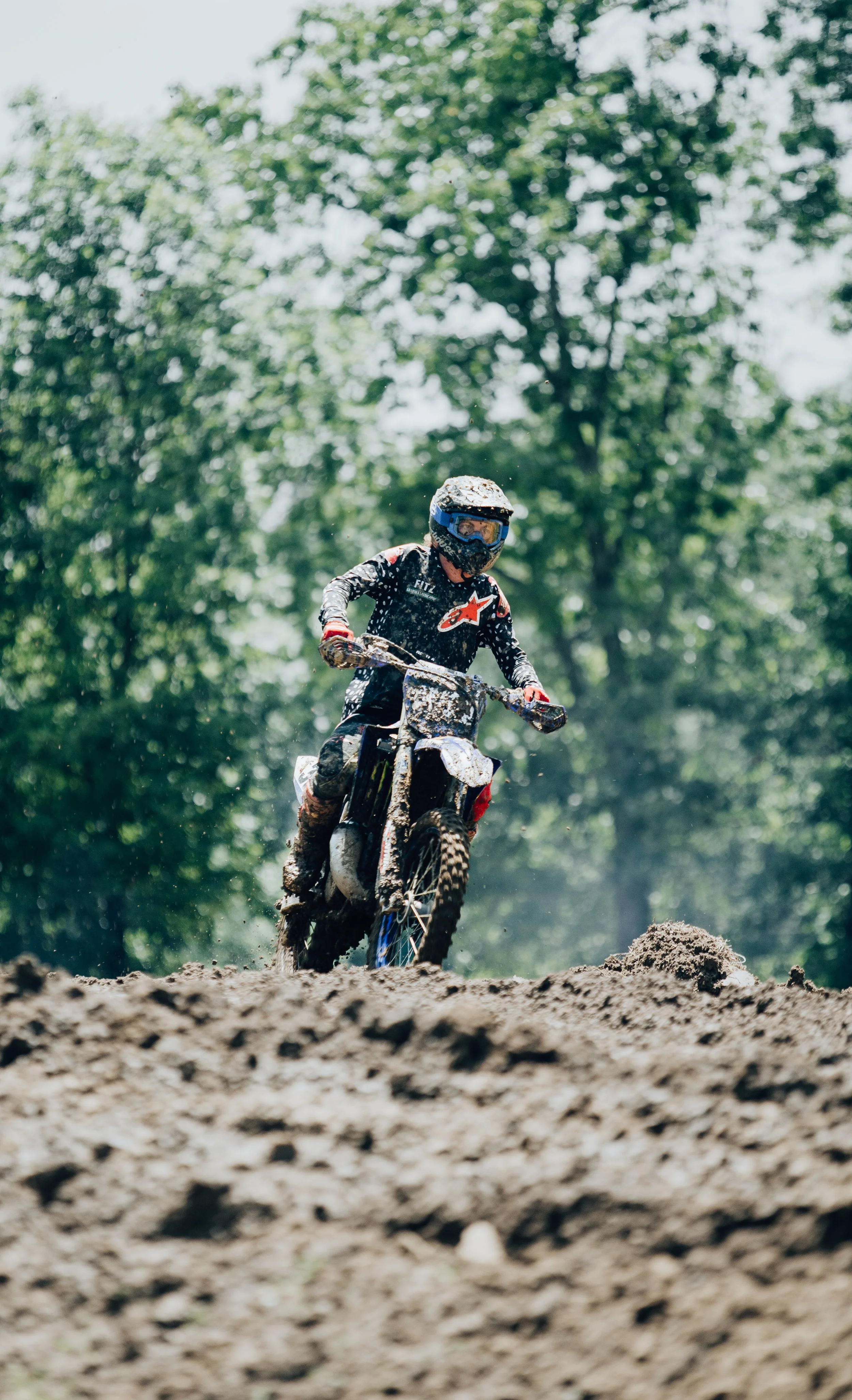 A person riding a dirt bike on a muddy trail surrounded by green trees, wearing a helmet and protective gear.