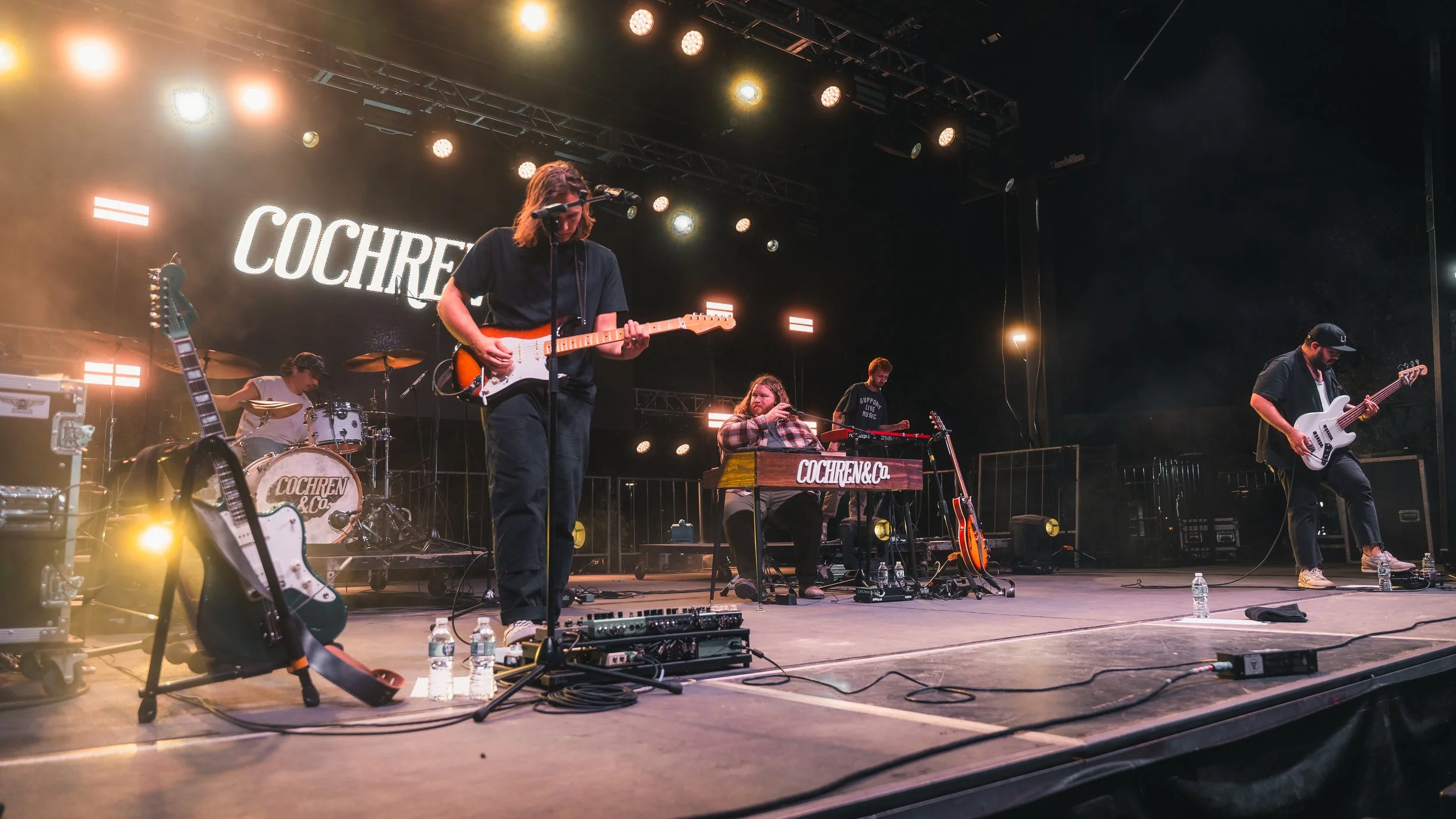 A band of five musicians performing live on stage, playing guitars, drums, keyboard, and singing into microphones. The stage background displays the words 'COCHREN & CO.' in large letters. The stage is lit with yellow and white lights, and water bottles are on the stage floor.