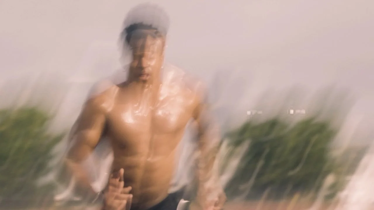 A shirtless man wearing a helmet running outdoors, with a sunset or sunrise sky in the background and some trees nearby.