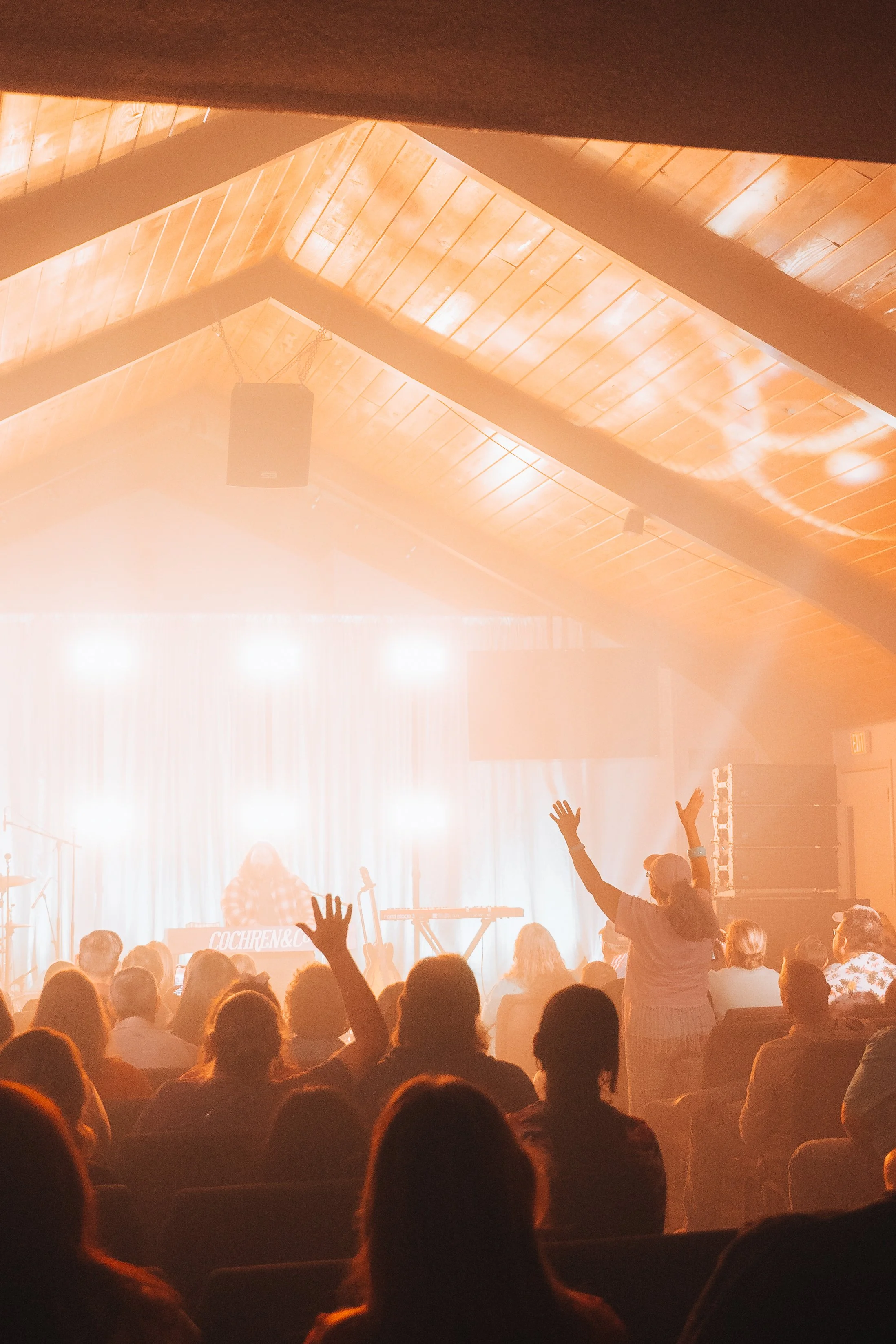 People sitting in an auditorium appear to enjoy a performance, some raising their hands, with stage lights illuminating a performer and musical instruments, all inside a wooden ceiling venue.