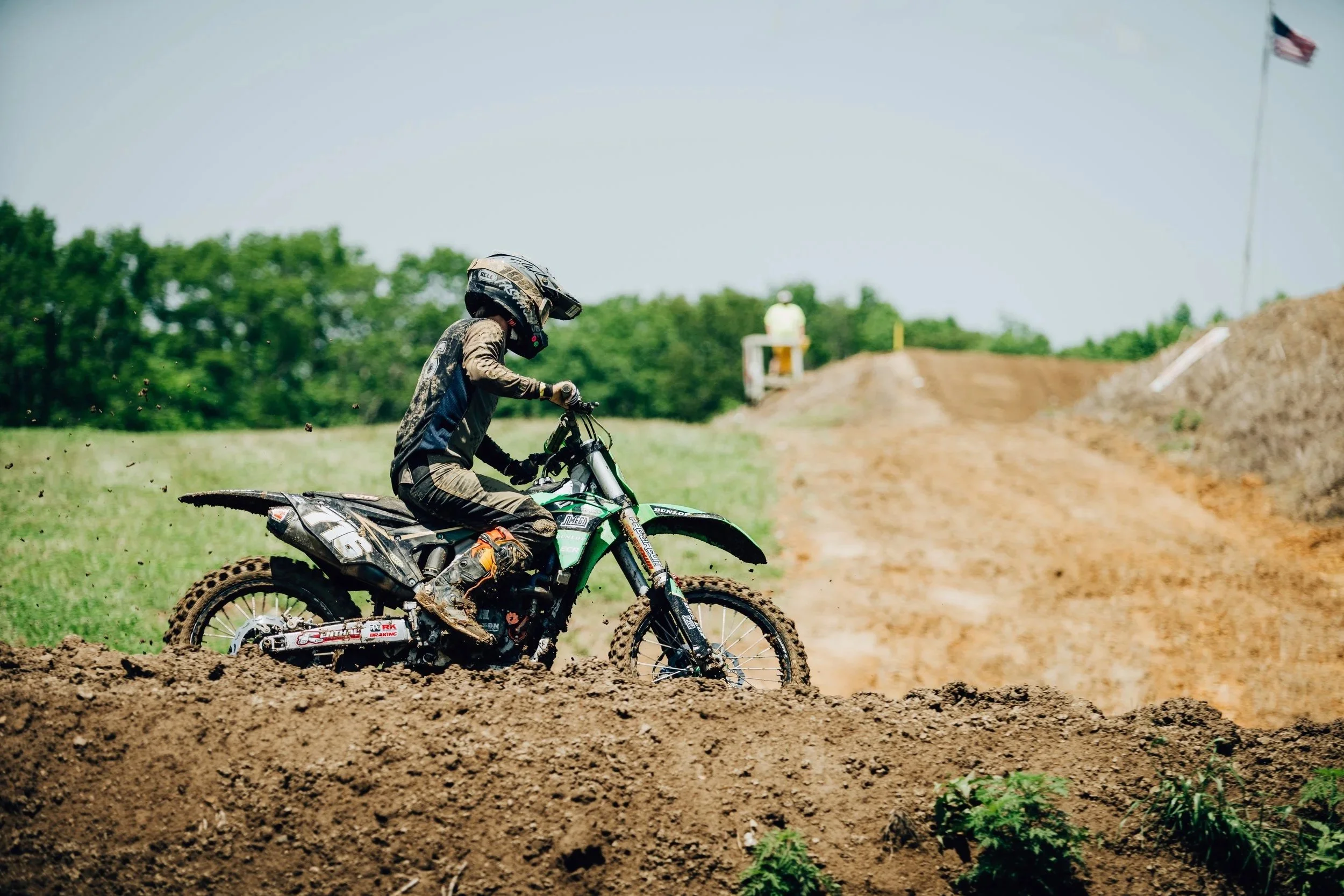 A person wearing protective gear riding a dirt bike on a motocross track with dirt mounds and a flag in the background.