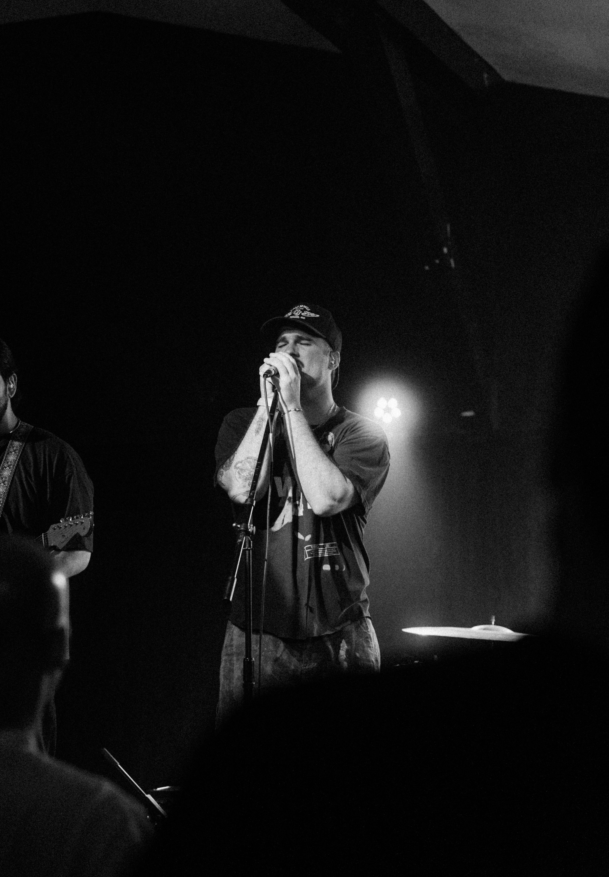A black and white photo of a male singer with tattoos on his arms, wearing a cap and casual clothing, performing on stage with a microphone in his hand and a drum set in the background.