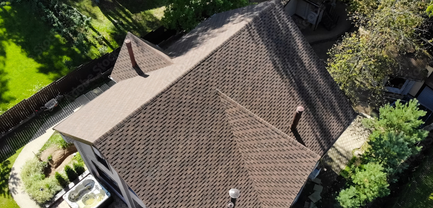 Aerial view of a house with a brown shingle roof, surrounded by green trees and a fenced backyard with a concrete patio and a hot tub.