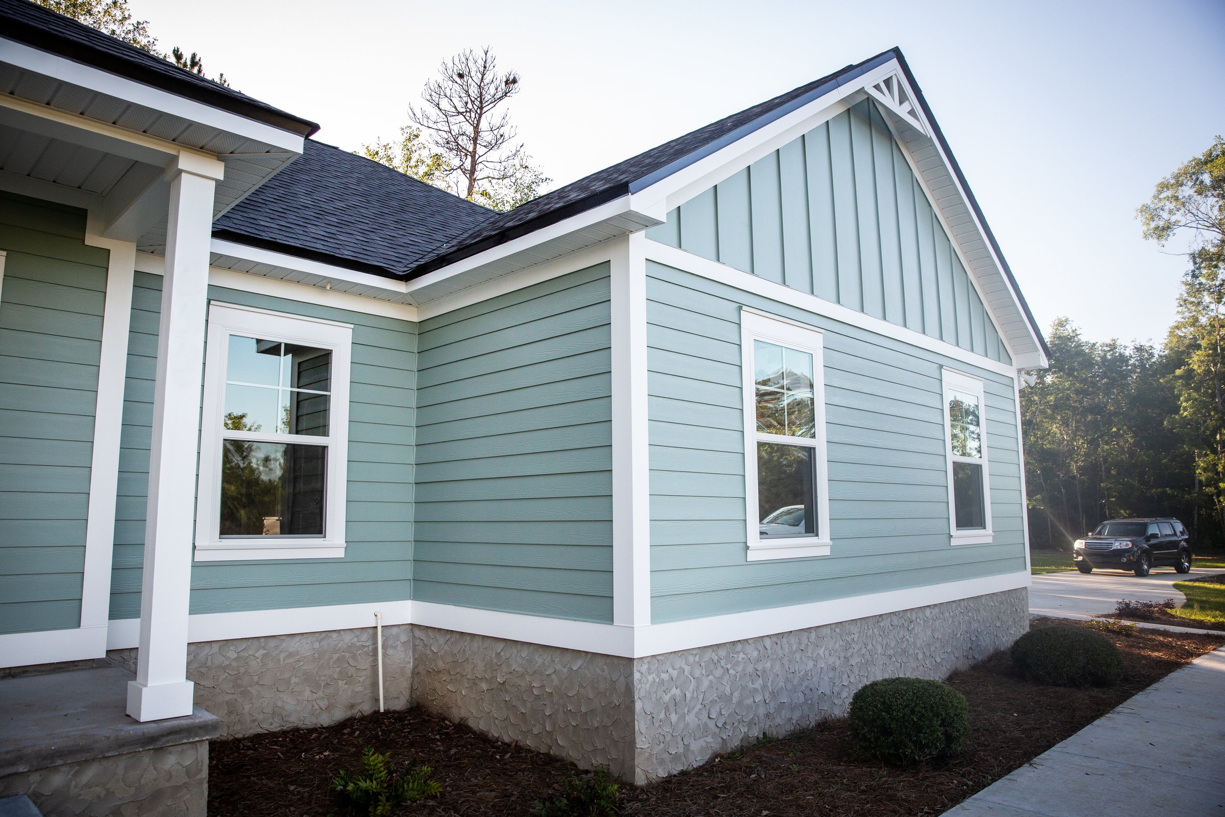 Side view of a light blue house with white trim, two white-framed windows, a stone foundation, and a black car parked nearby. Tall trees are in the background.