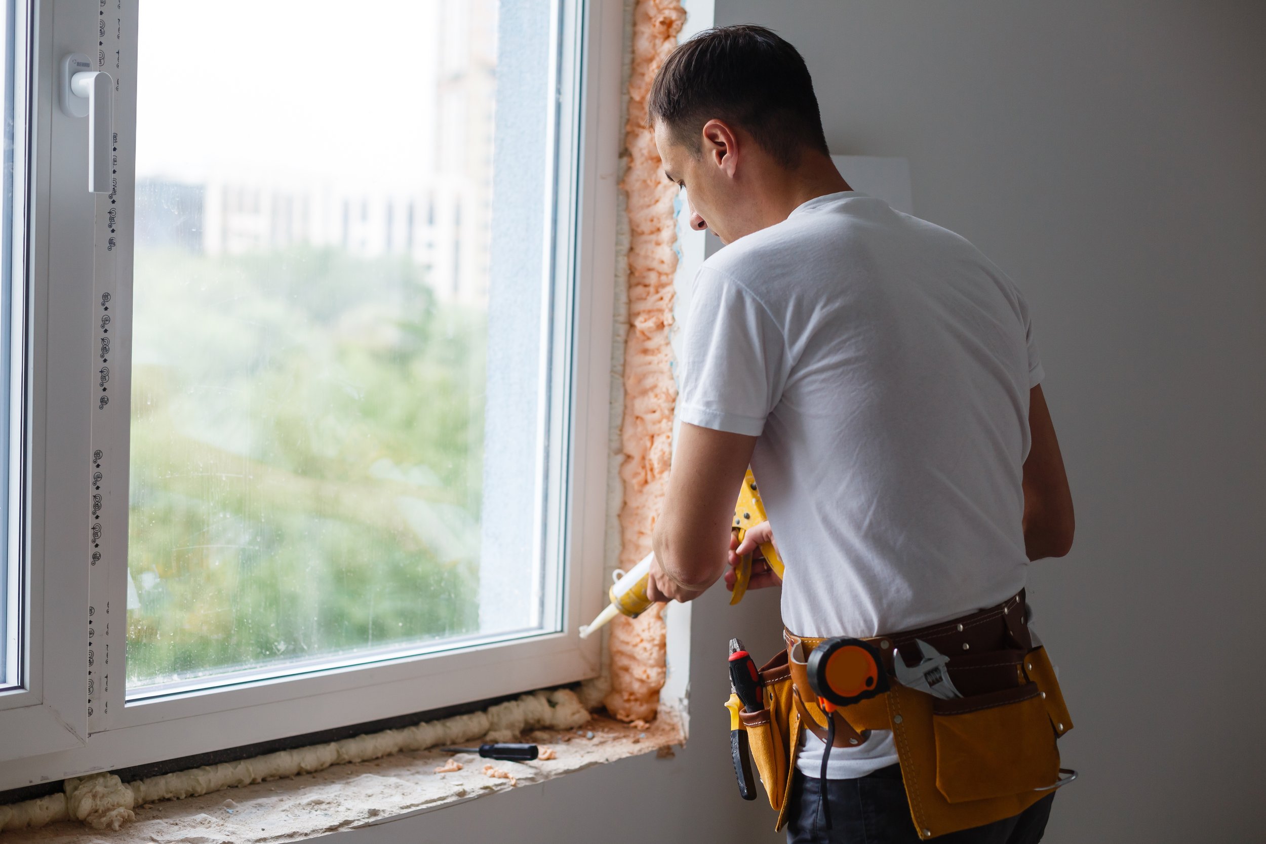 A man in a white t-shirt and tool belt working on a window installation or repair inside a house, with exposed insulation around the window frame.