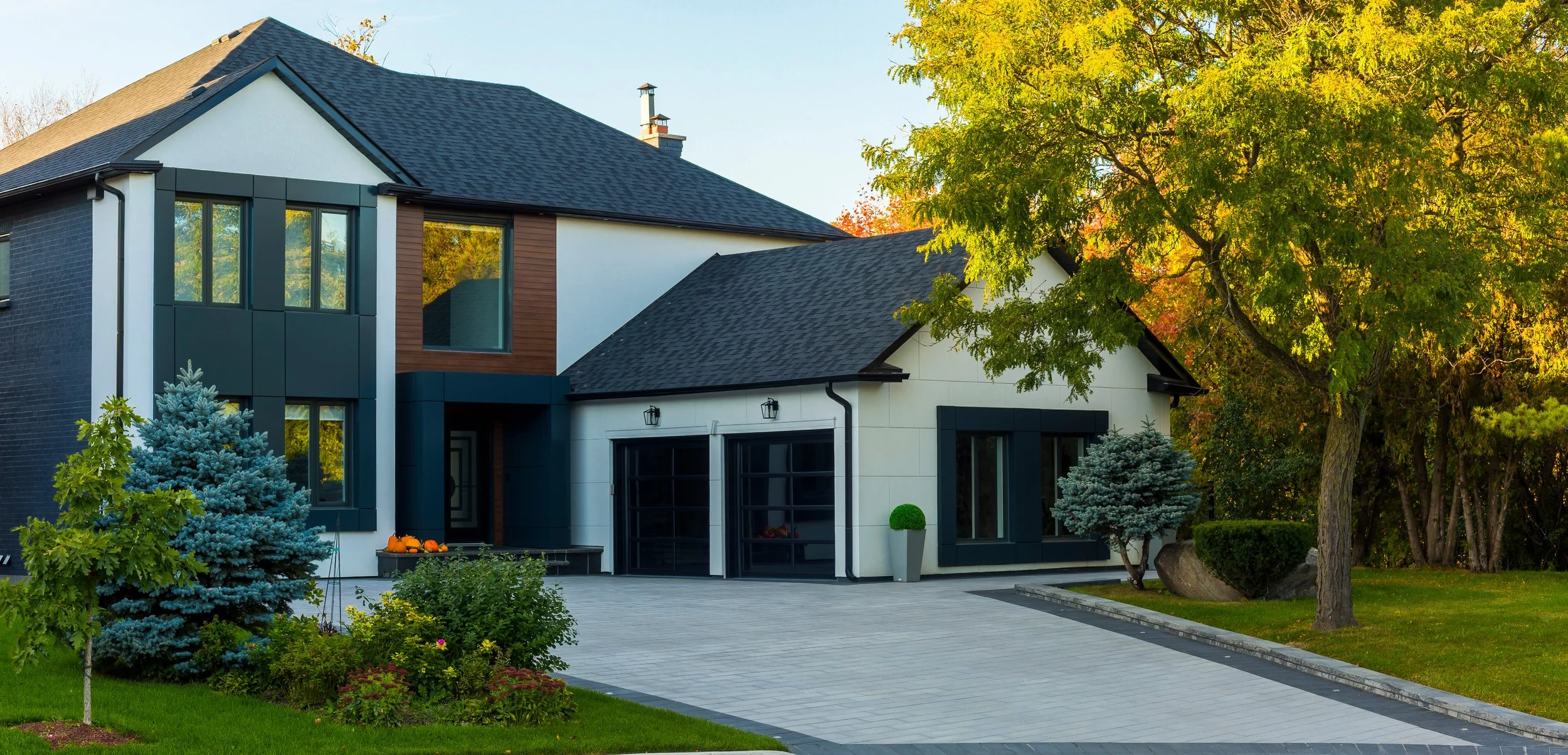 Modern two-story house with white and dark gray exterior, large windows, a black roof, surrounded by green trees and shrubs, with a paved driveway and front garden.