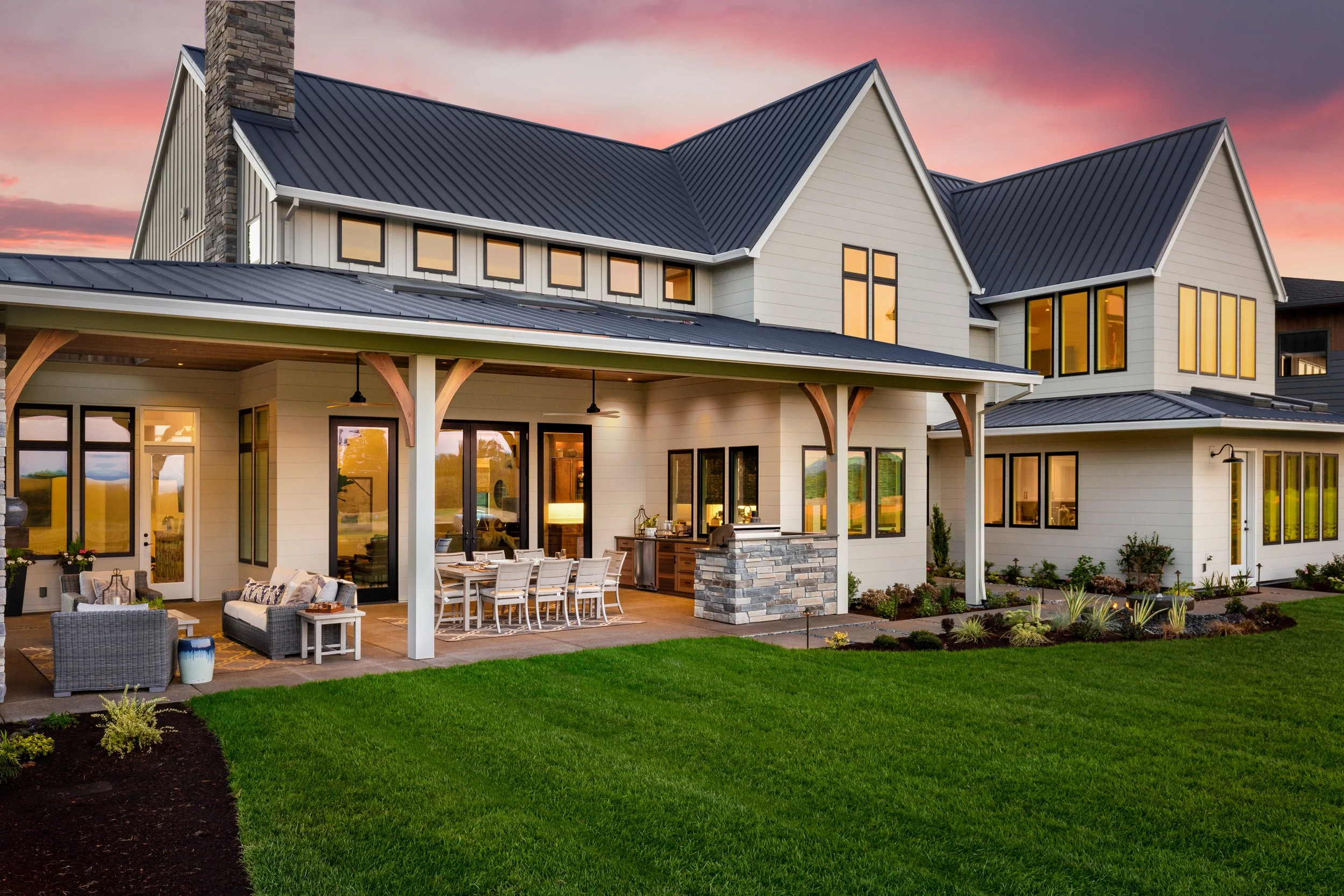 A modern two-story house with beige exterior walls, black window frames, and a metal roof, set against a sunset sky. The house has a spacious backyard with a patio area furnished with outdoor seating, a dining table, and an outdoor kitchen. Andersen