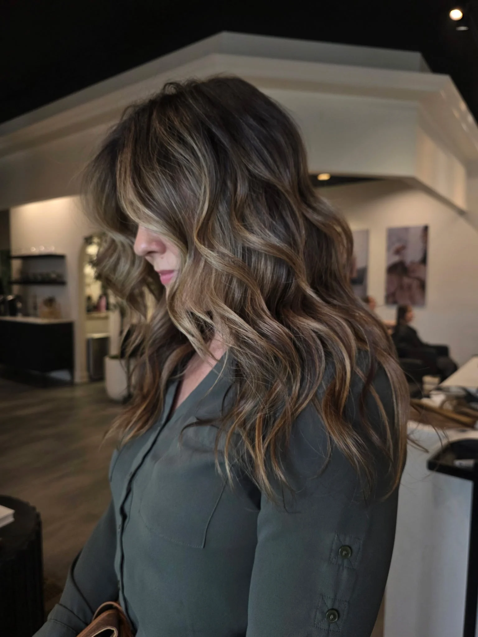 Profile of a woman with wavy, highlighted brown hair in a salon or similar setting.