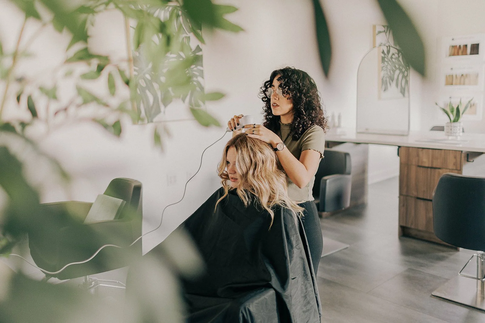 Hair stylist using a tool on a woman's hair in a salon, with modern decor and plants.